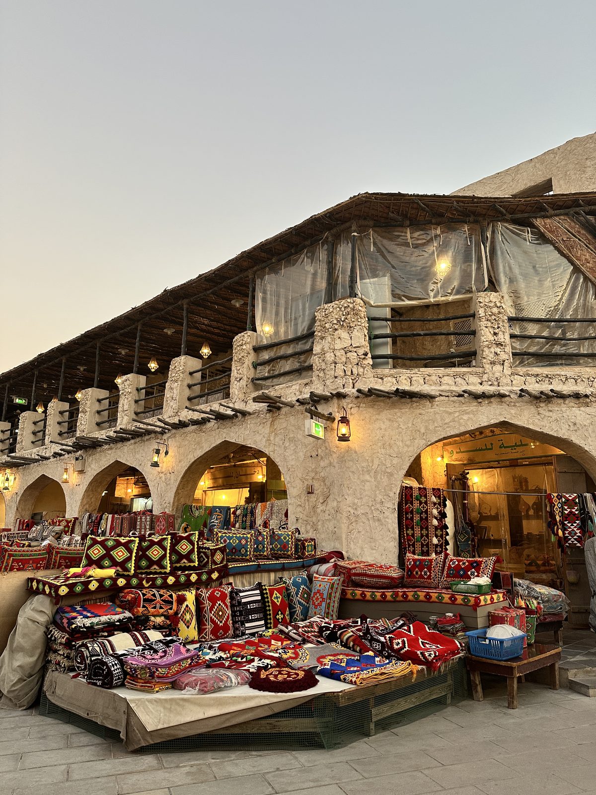 A mud-brick arcade in Souq Waqif at dusk with carpets and embroidered cushions spread out under arches strung with lanterns