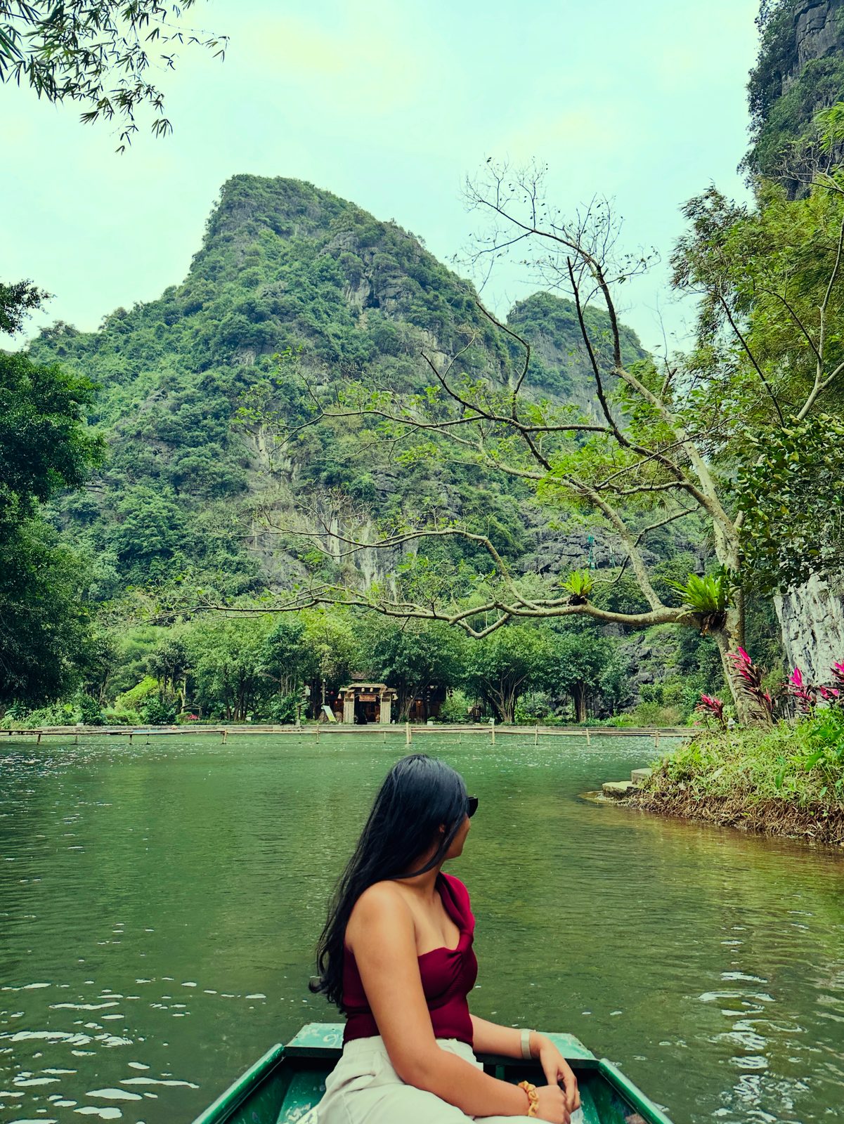 A woman sitting at the bow of a rowing boat on a green river at Tam Coc, a karst peak rising ahead