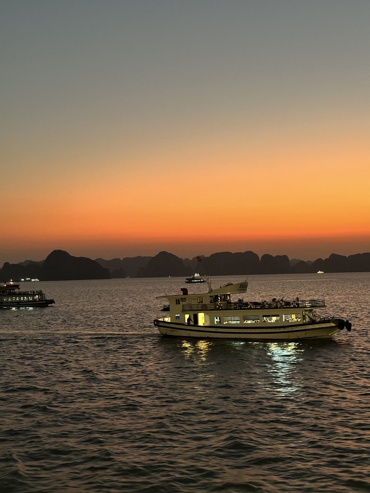 Sunset over Ha Long Bay, karst islands in silhouette and a lit cruise boat in the foreground