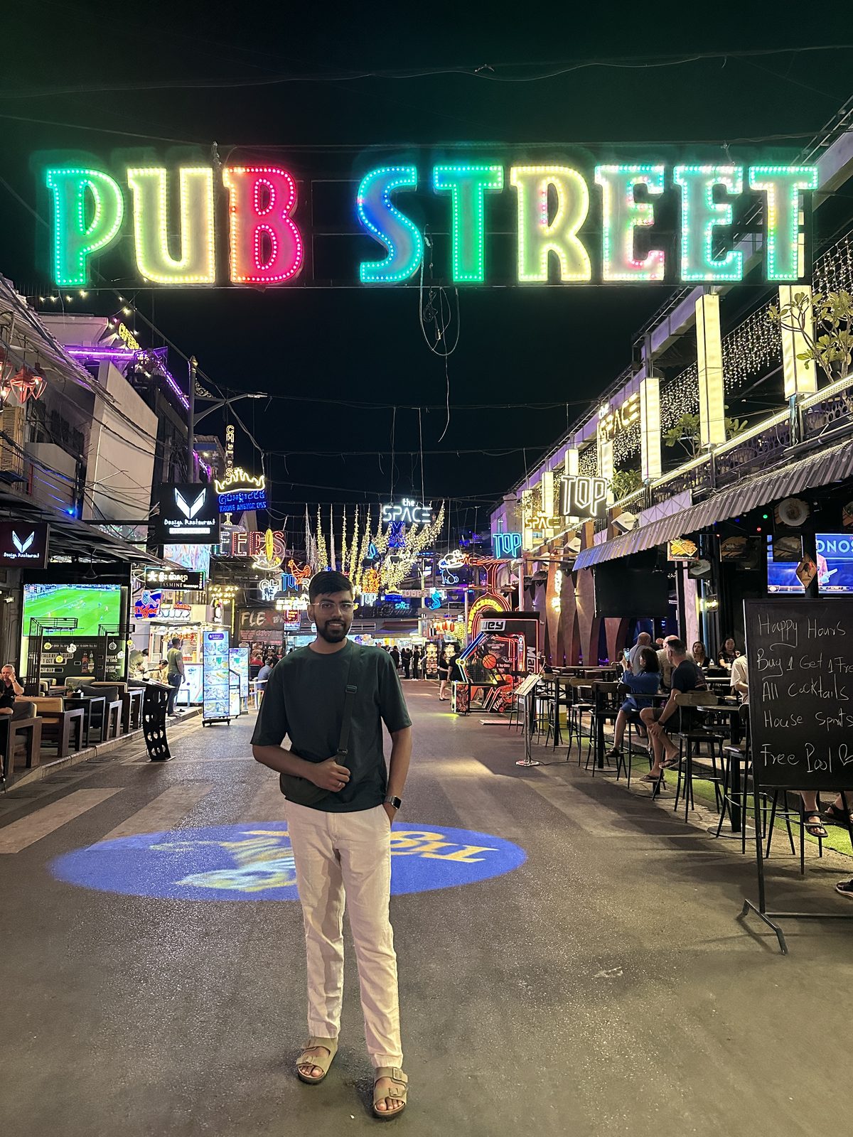 A man standing under the neon Pub Street sign in Siem Reap at night, bars and lights either side