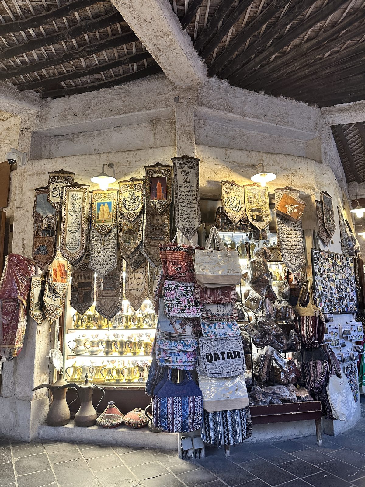 A souq shop hung with embroidered wall hangings above a lit cabinet of brass dallah coffee pots