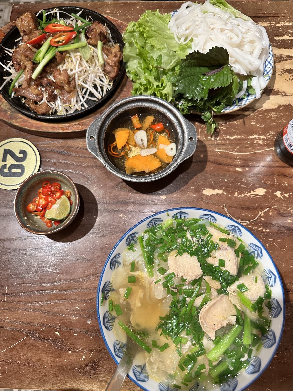 A Hanoi table with sizzling pork and bean sprouts, rice noodles, a plate of herbs, and a bowl of chicken noodle soup