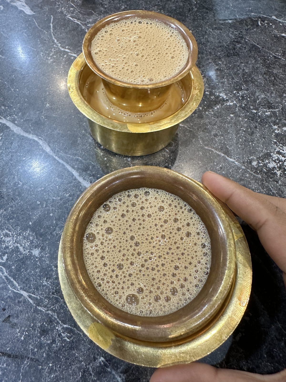Two brass davara-tumbler sets of South Indian filter coffee on a dark marble counter, one being poured and frothed