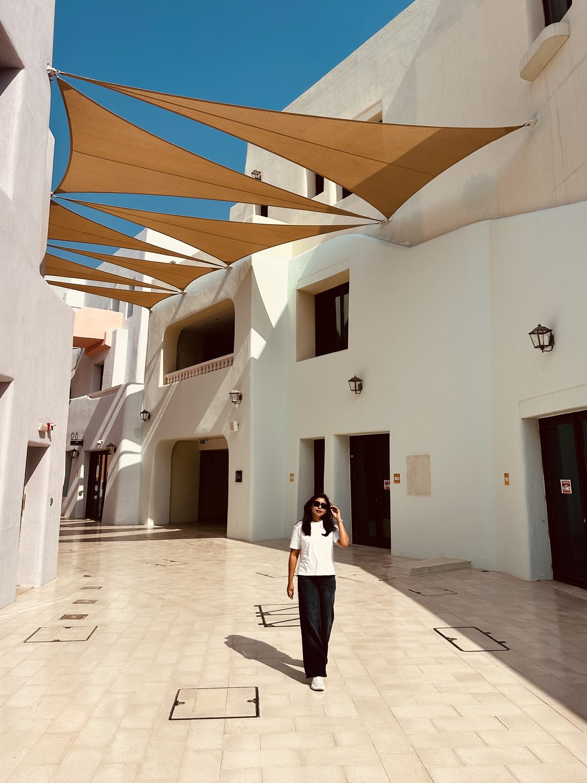 A woman walking through an empty plaza of white modernist buildings under triangular sand-coloured shade sails