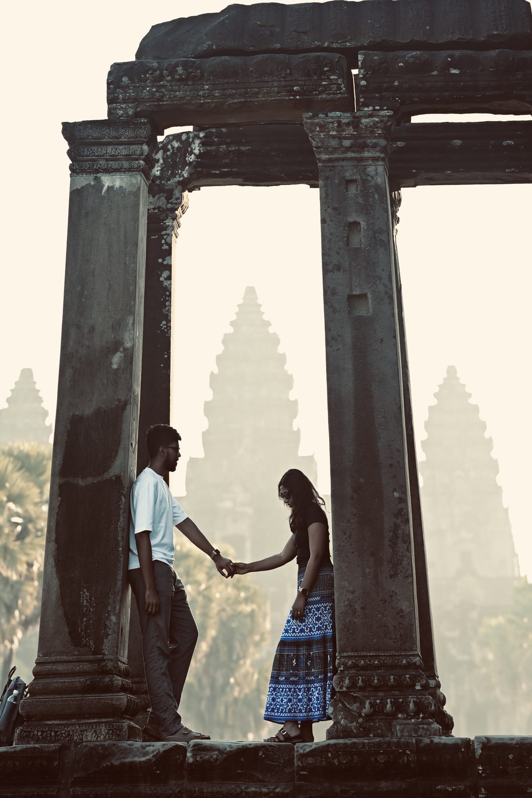 A couple holding hands between two stone columns, with the towers of Angkor Wat hazy in the light behind