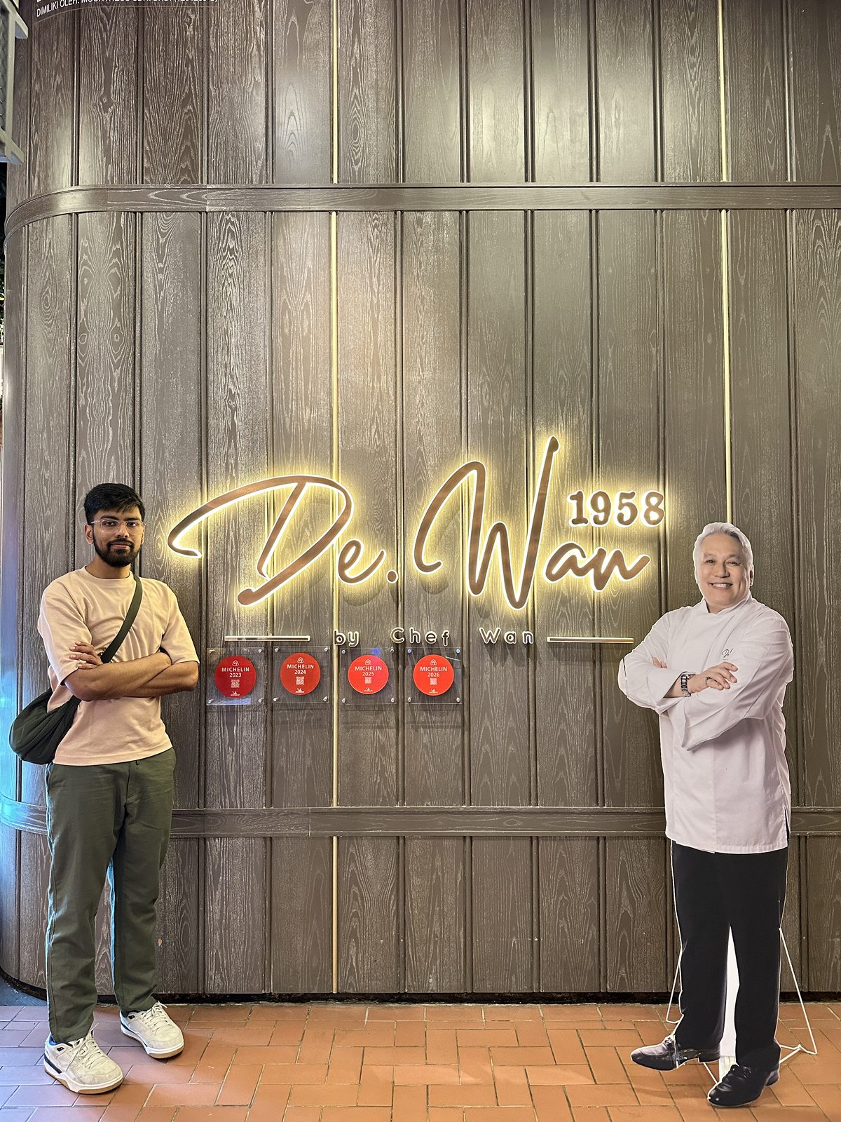 A man standing beside the illuminated De.Wan 1958 sign, with four Michelin plaques mounted below it and a life-size cardboard cutout of Chef Wan alongside