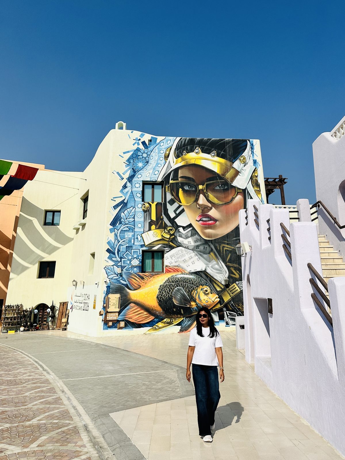 A woman walking past a huge mural of a woman in a keffiyeh and gold sunglasses above a painted fish, on the side of a white building