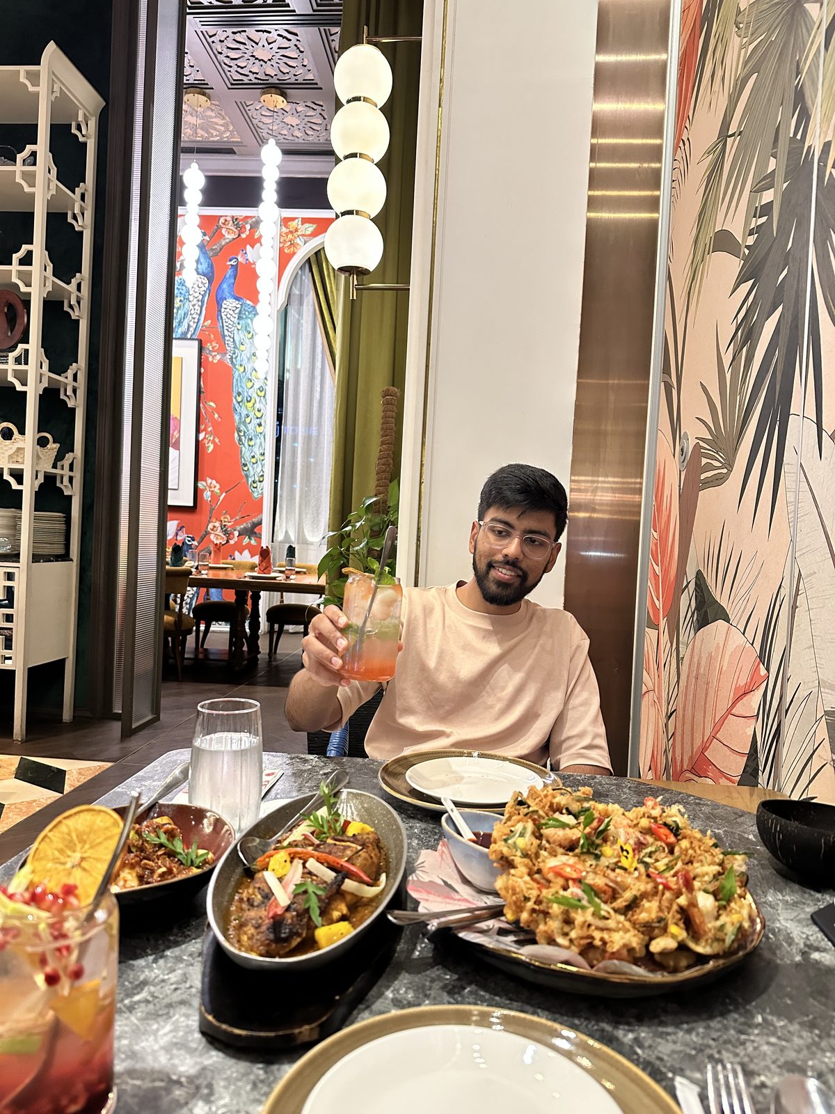 A man at a restaurant table raising a cocktail, with dishes of rendang, grilled fish and a prawn dish in front of him