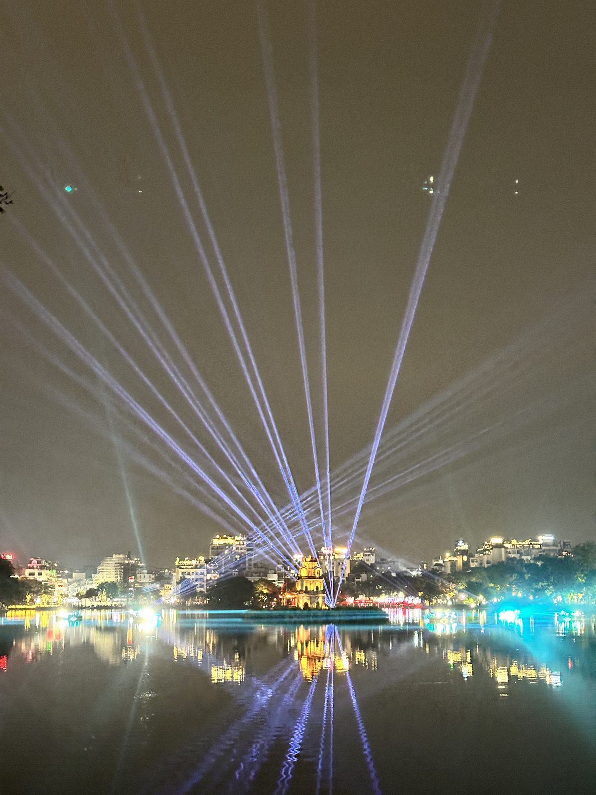 A laser show over Hoan Kiem Lake in Hanoi at night, beams fanning out over the water with the Turtle Tower lit below