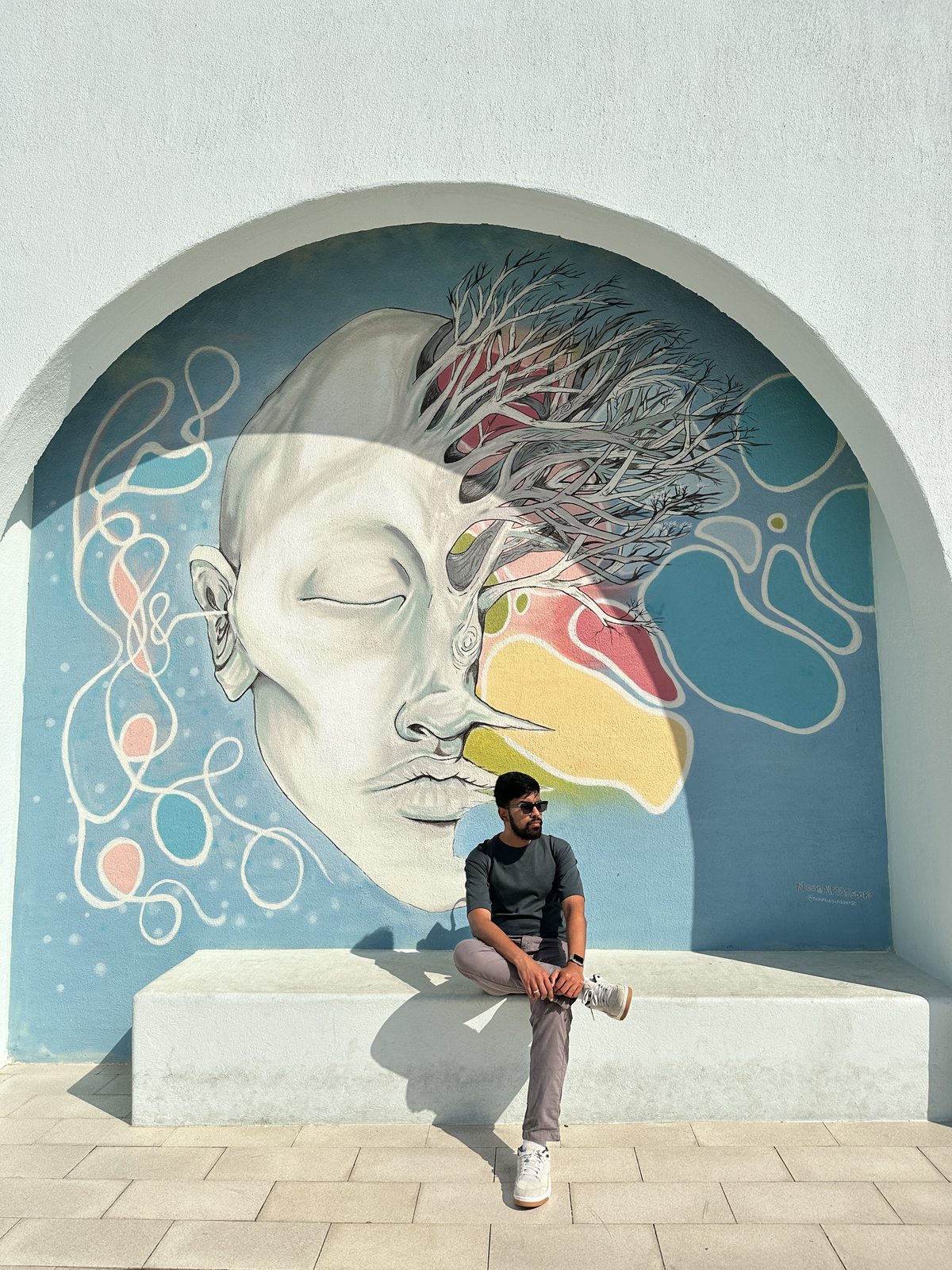 A man sitting beneath a large mural of a sleeping face with tree branches growing from it, painted inside an arched recess