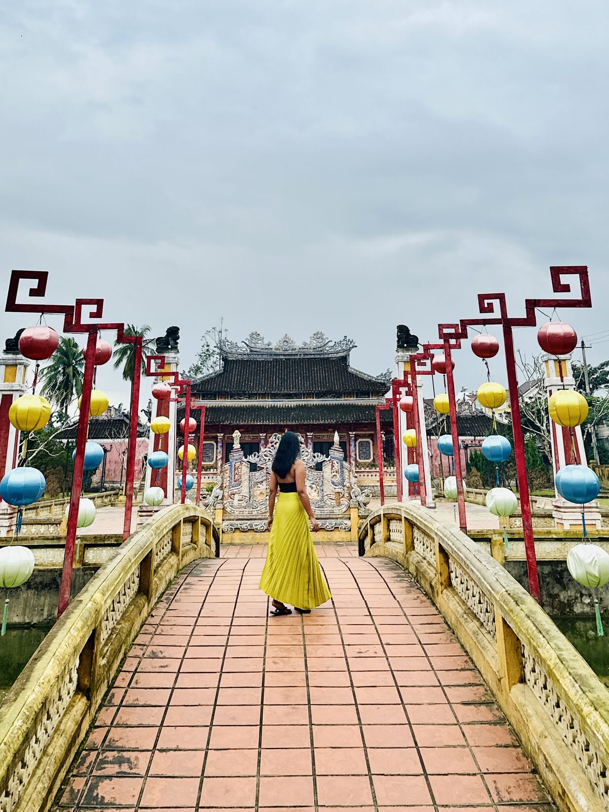 A woman on an arched bridge lined with red frames and coloured paper lanterns, leading to an ornate temple