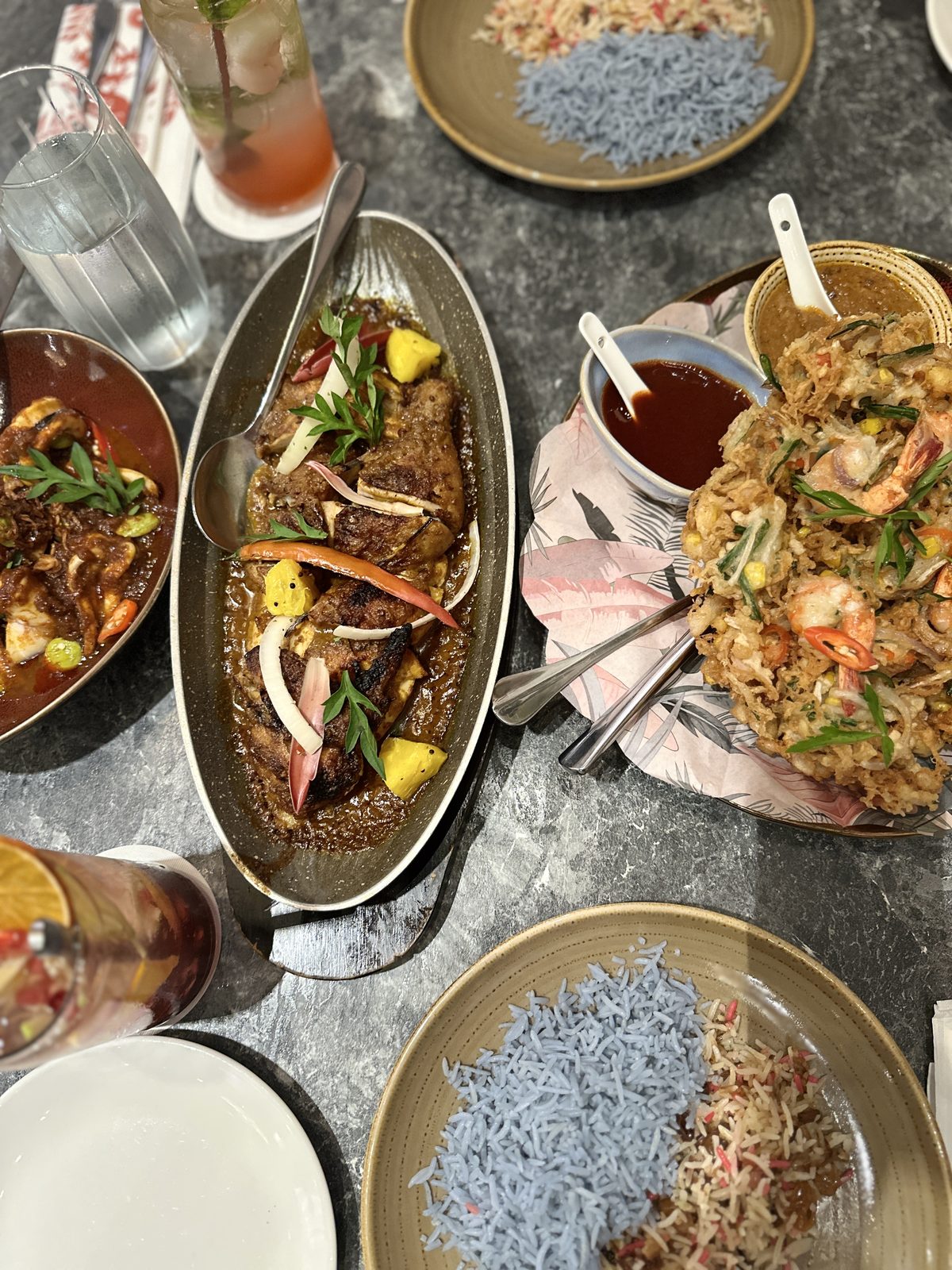 An overhead view of a Malaysian table: nasi kerabu with pale blue rice, beef rendang, grilled fish in curry, and a prawn dish