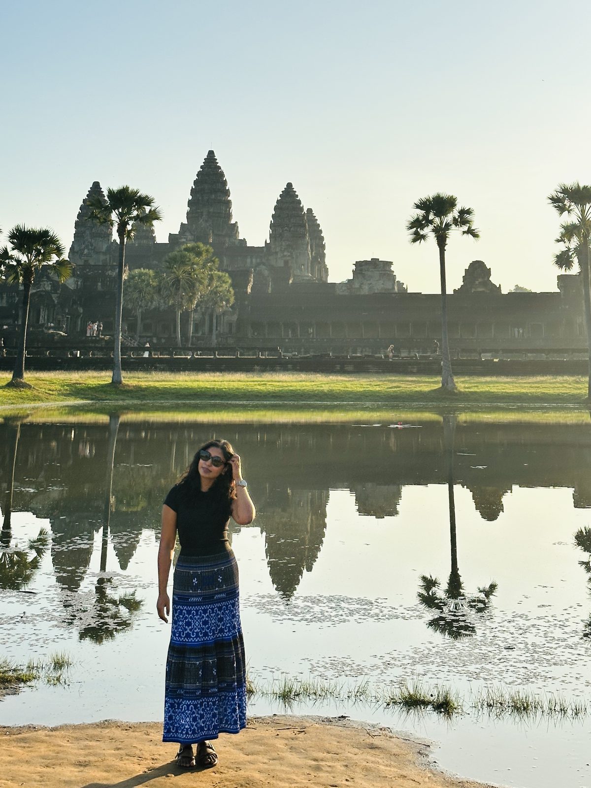 A woman standing in front of Angkor Wat and its reflecting pool in full morning sunlight, palm trees around