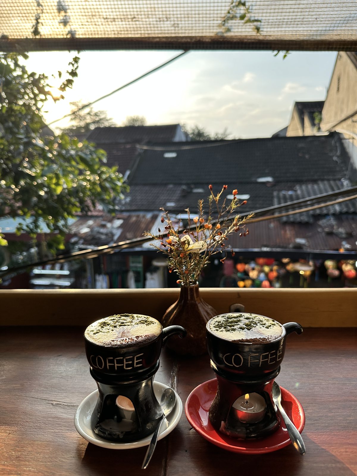 Two Vietnamese egg coffees on candle warmers at a window overlooking Hoi An rooftops at golden hour