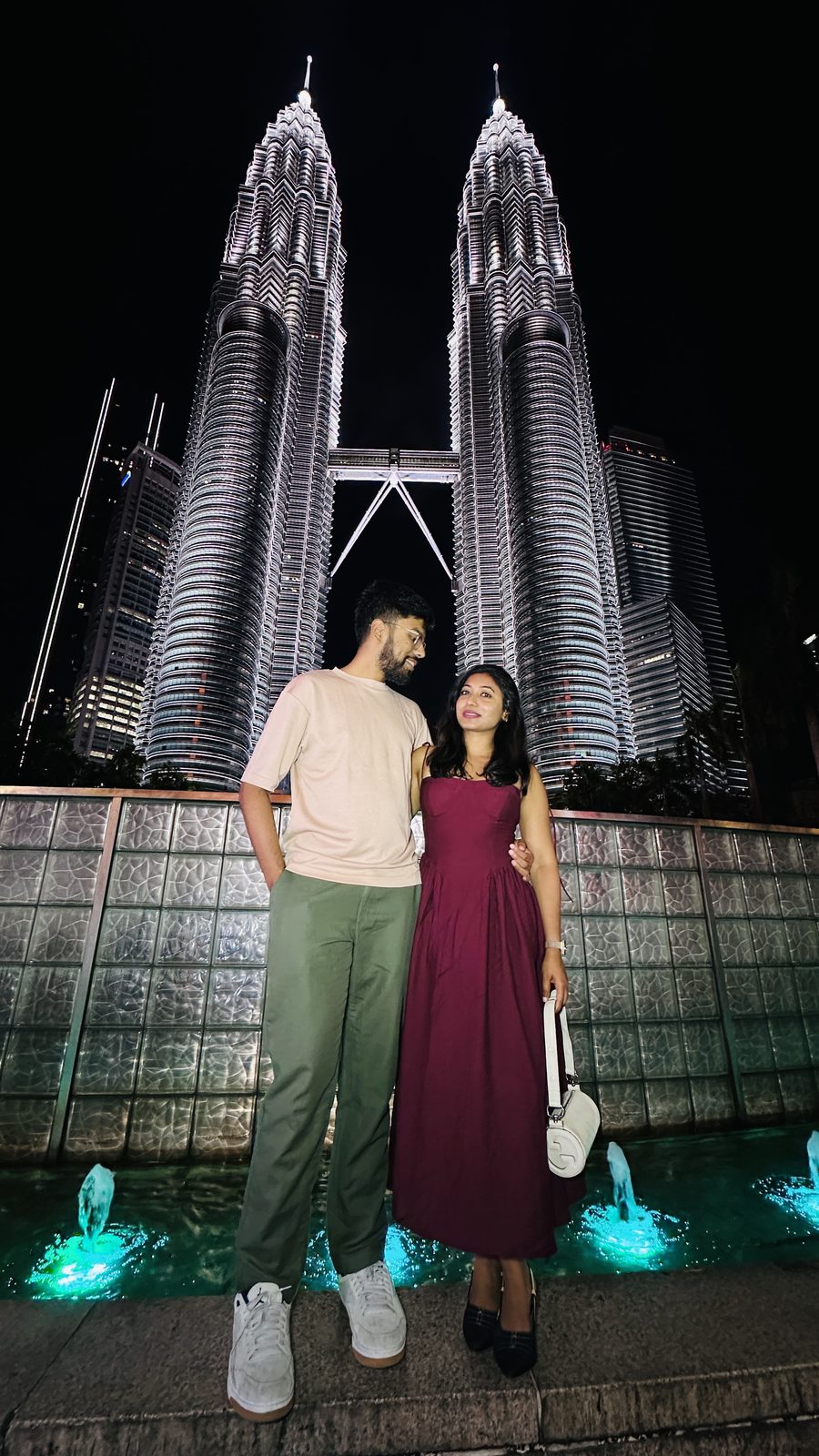 A couple standing in front of the illuminated Petronas Twin Towers at night, with lit fountains behind them