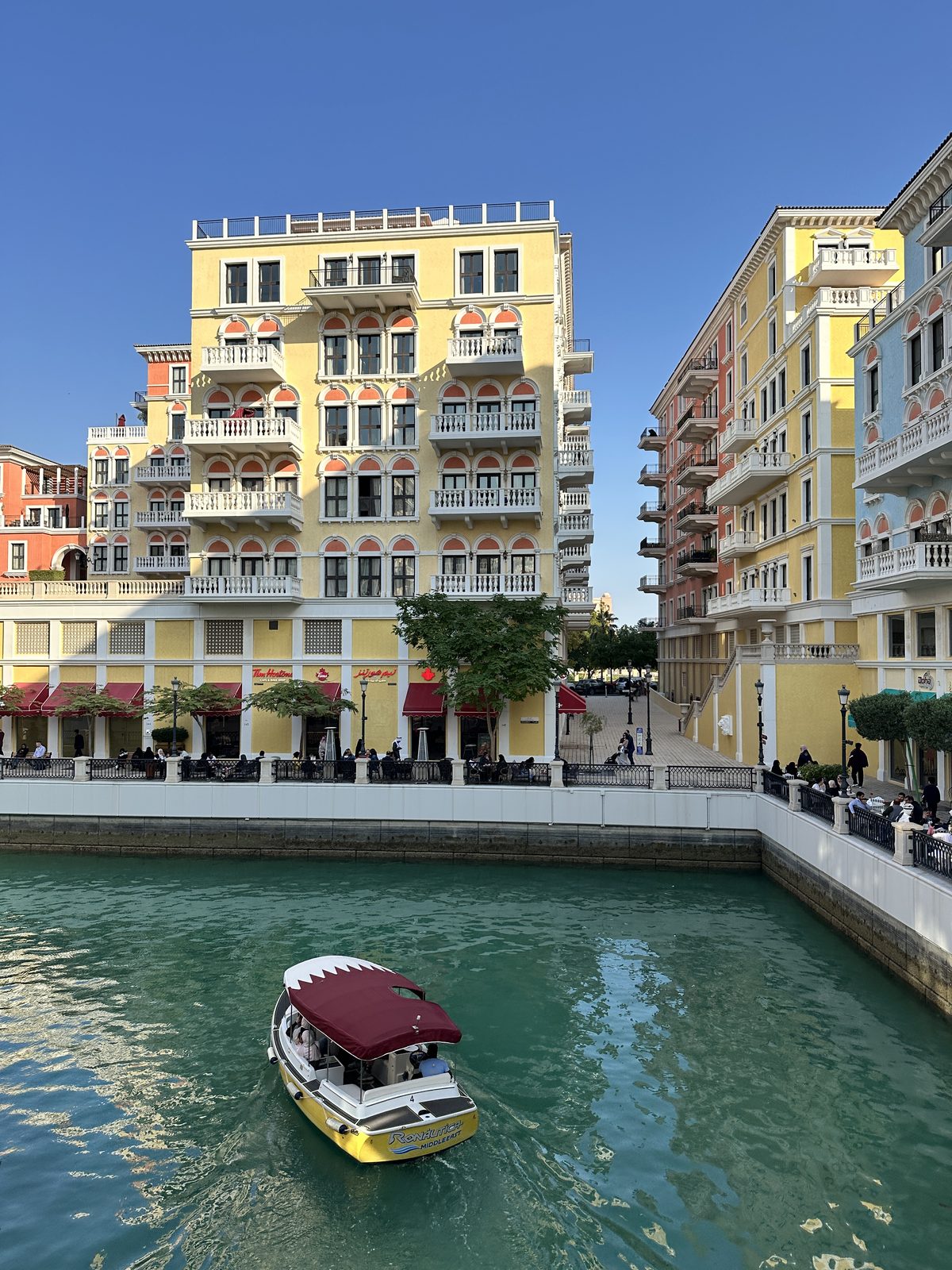 A canal at The Pearl lined with pastel yellow and orange Venetian-style buildings, with a small water taxi under a maroon Qatari-flag canopy