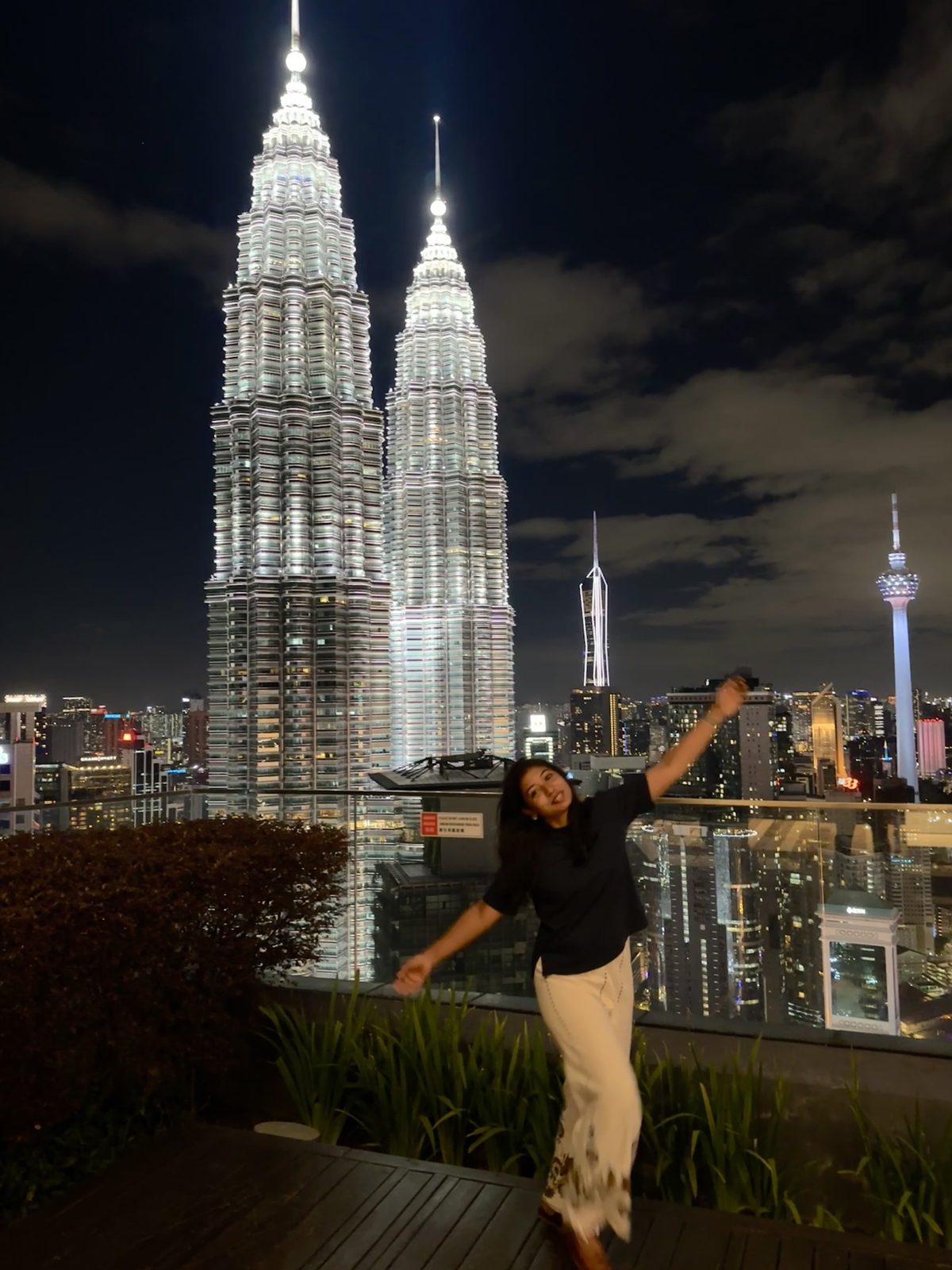 A woman on a rooftop terrace at night, arm raised mid-movement, with the lit Petronas Towers and the whole Kuala Lumpur skyline behind her