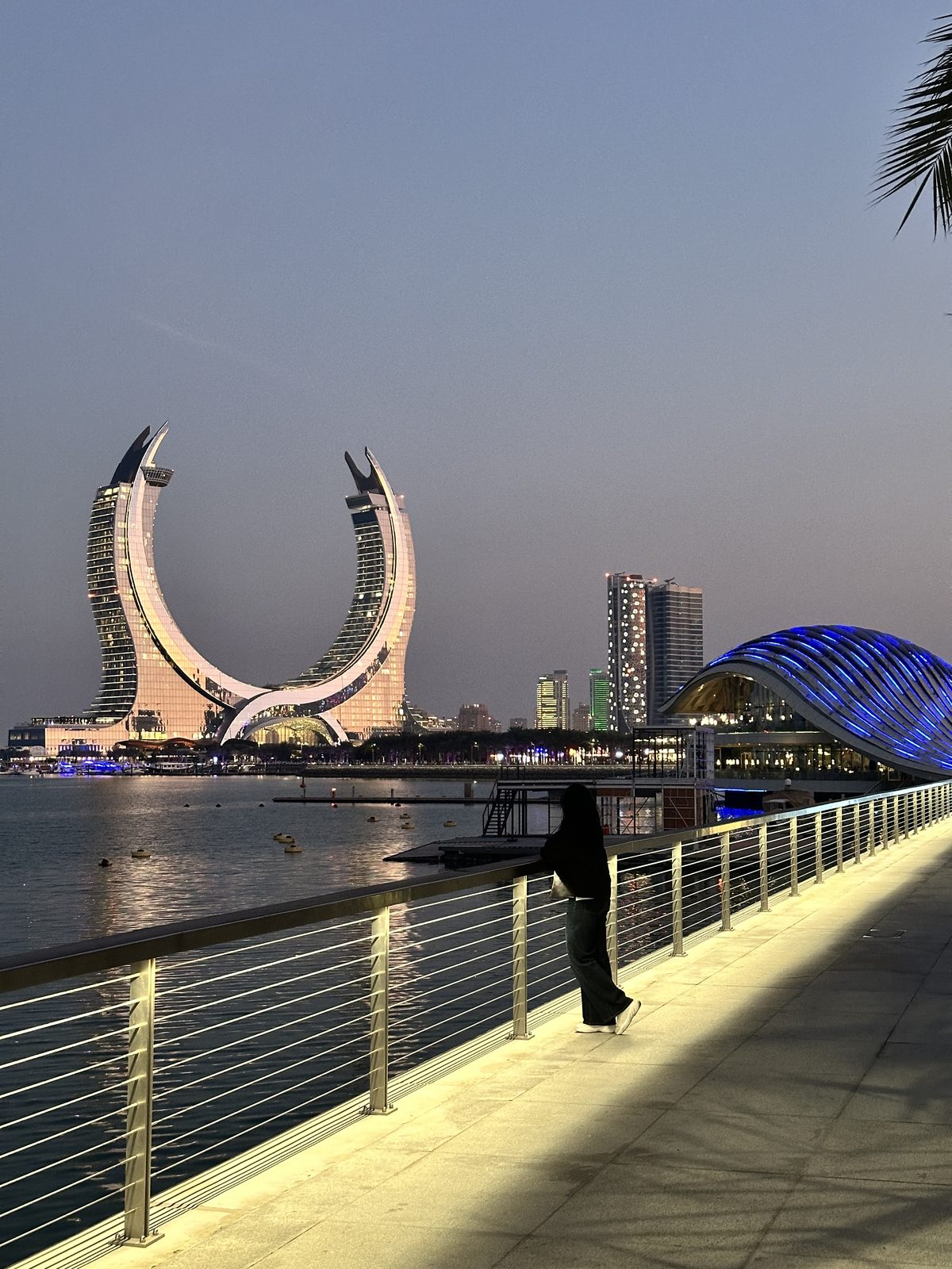A woman at a marina railing at dusk looking across the water at the crescent-shaped Katara Towers, lit pink against a pale sky
