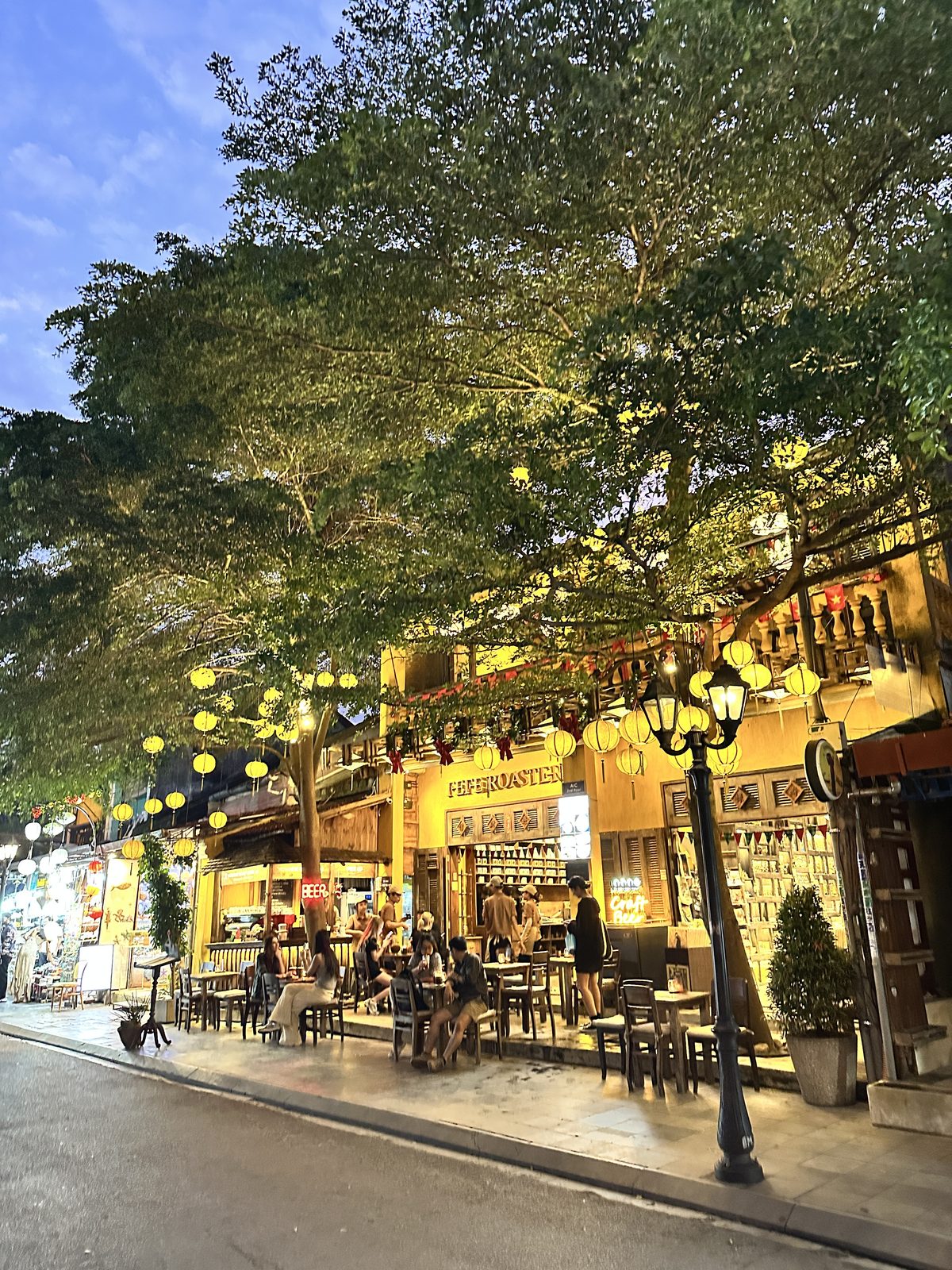A street in Hoi An at blue hour, silk lanterns strung through a large tree above a yellow cafe