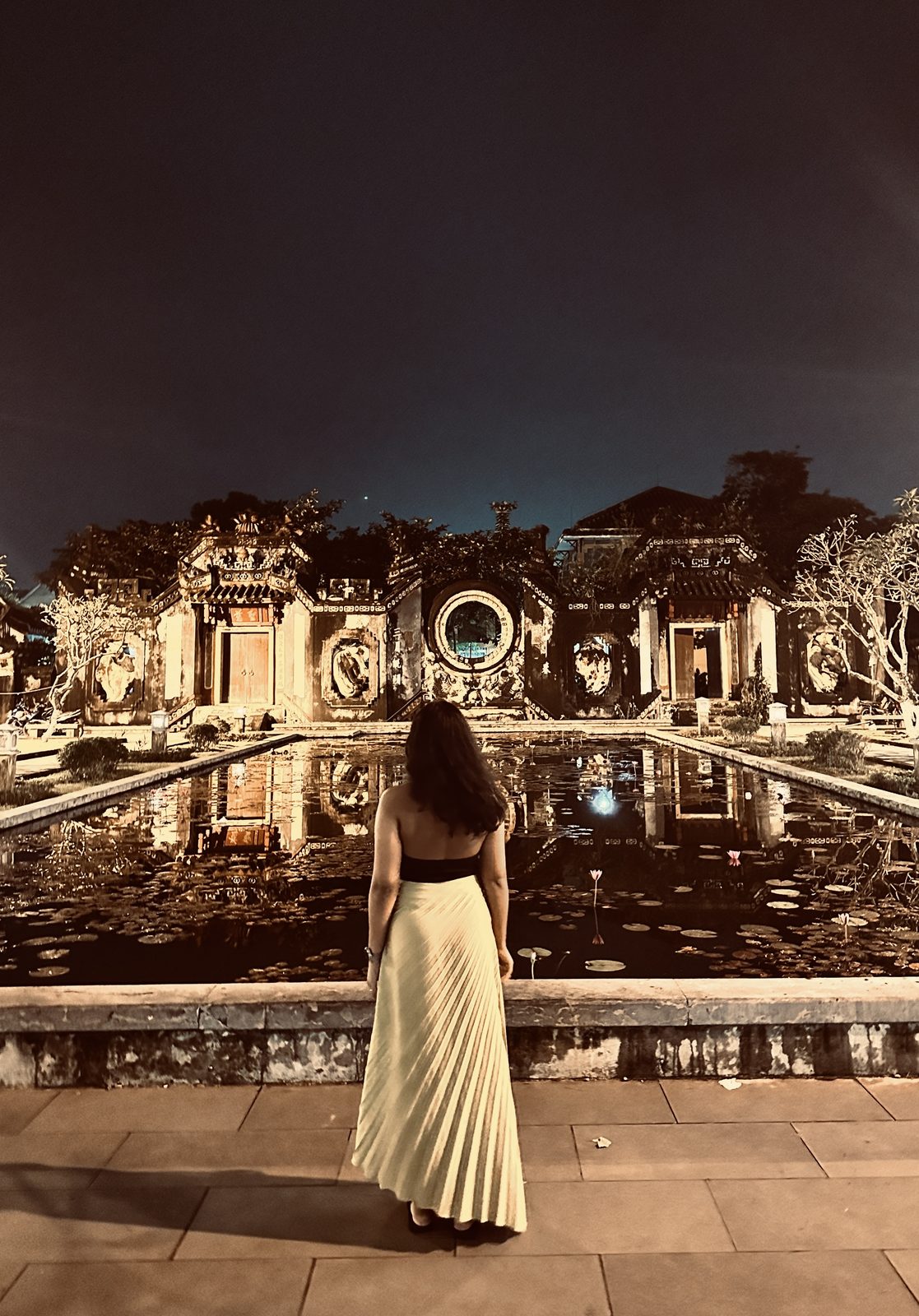 A woman in a cream skirt in front of an ornate old building and lily pond, lit warmly at night