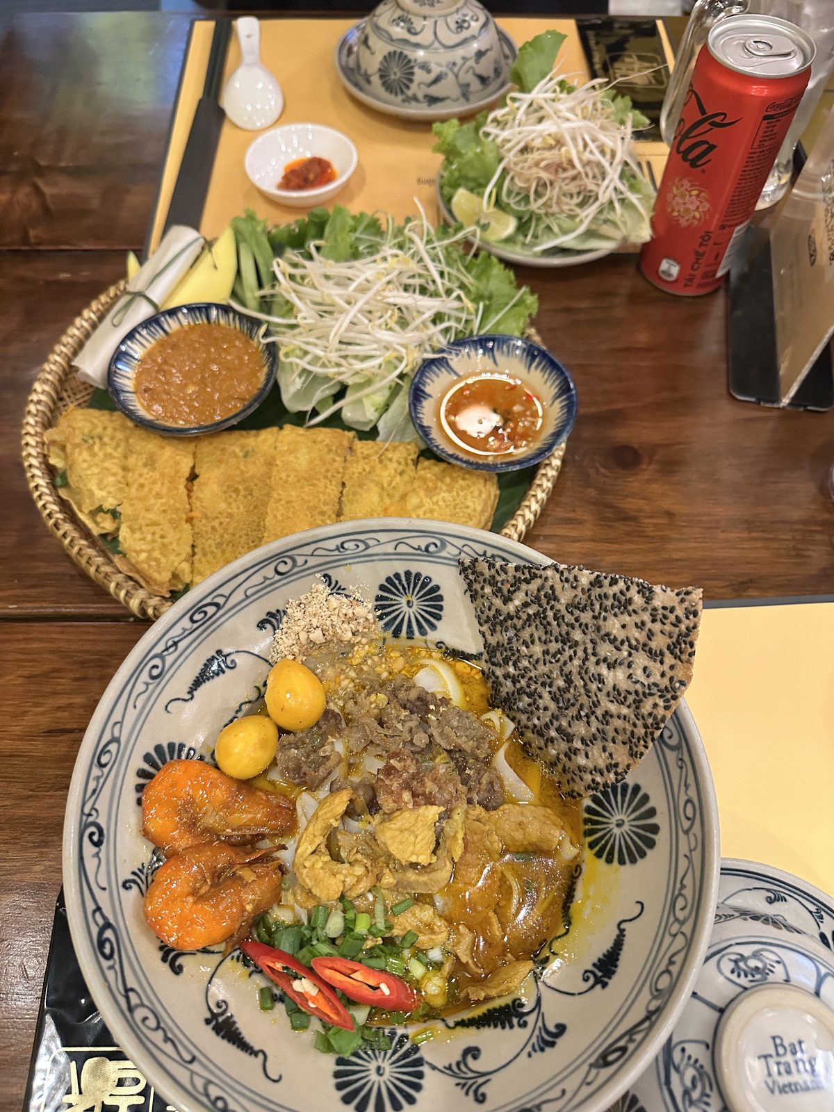 A Da Nang table with banh xeo crepes, herbs, peanut sauce, and a bowl of mi Quang with prawns, pork and a black sesame rice cracker