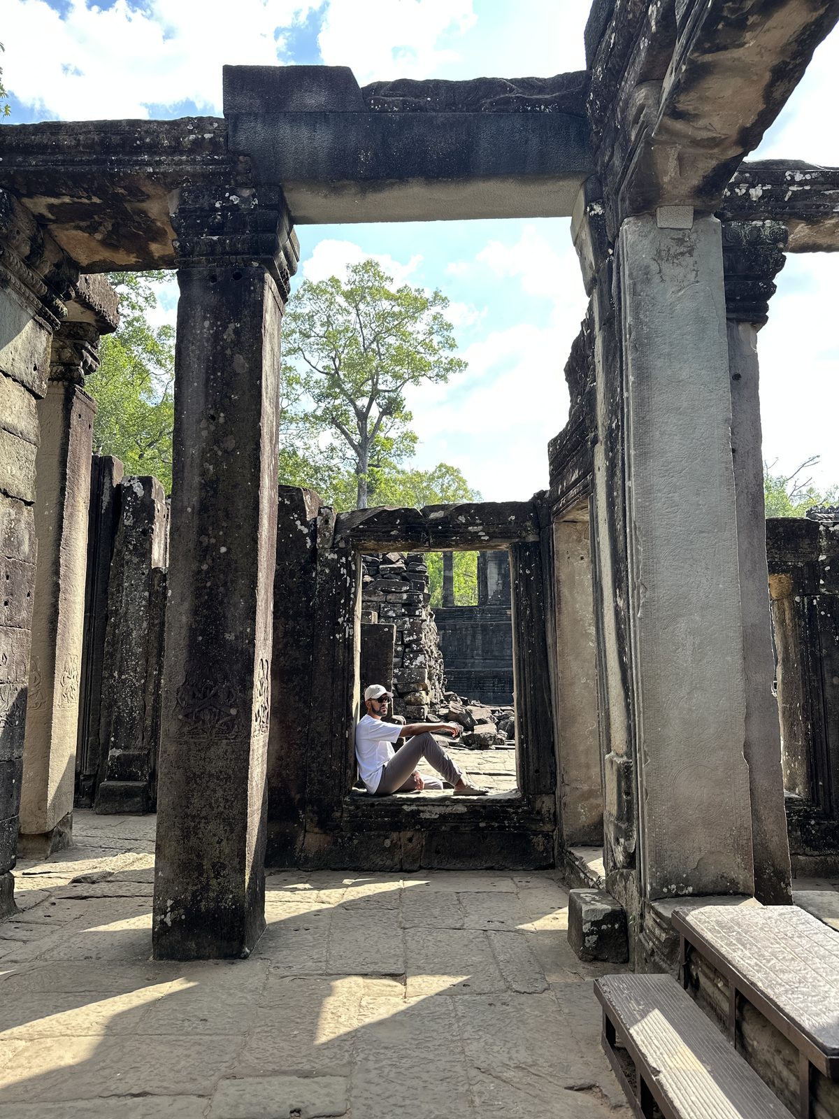A man sitting in a stone window frame at Bayon, framed by carved columns and bright green trees beyond