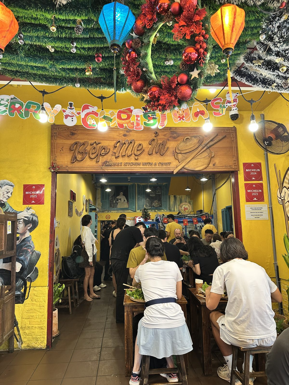 The entrance to Bep Me In in Saigon, with a carved wooden sign, three Michelin plaques and Christmas decorations