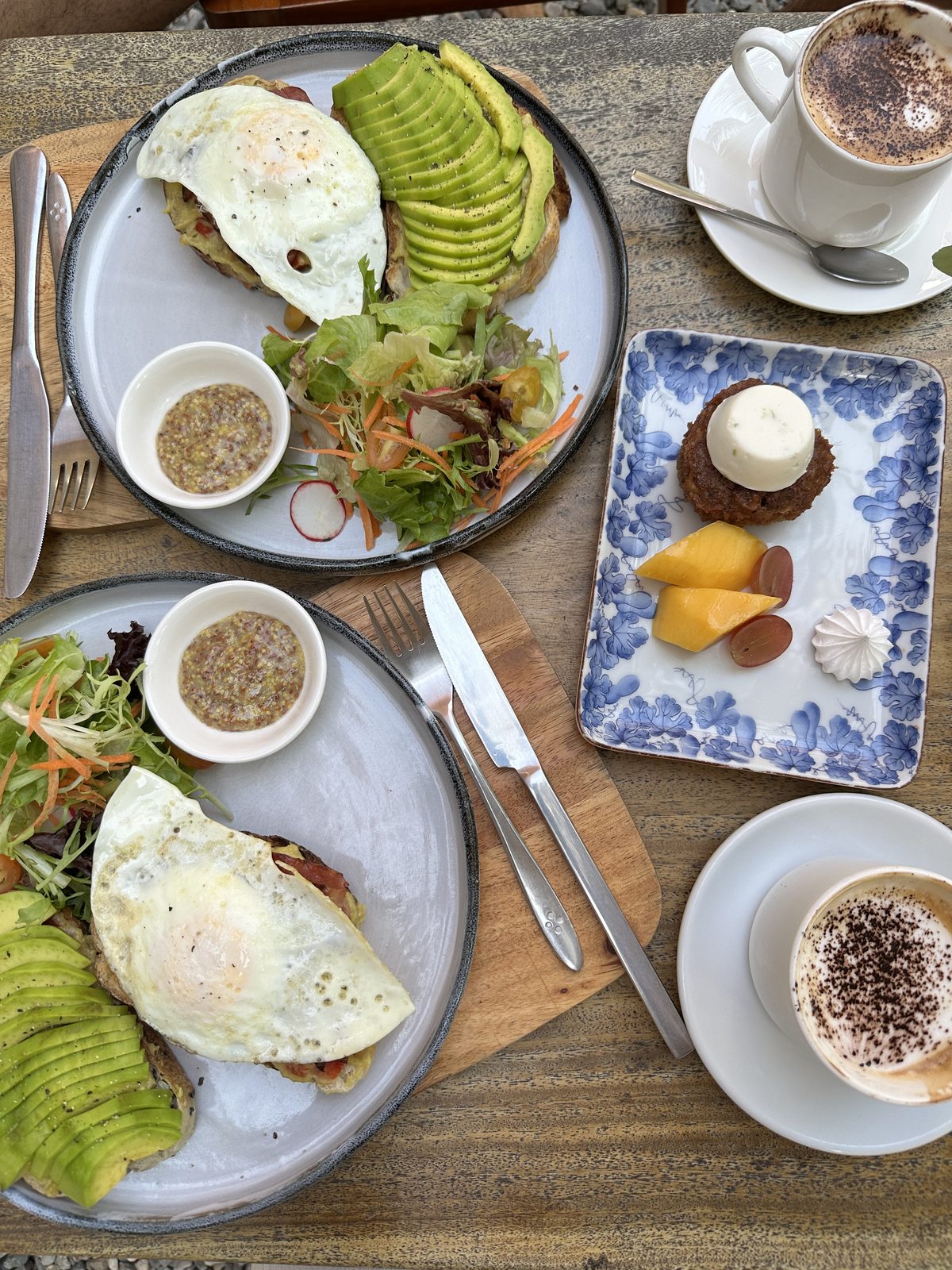 A brunch table with eggs and avocado on toast, salad, two cappuccinos and a small cake
