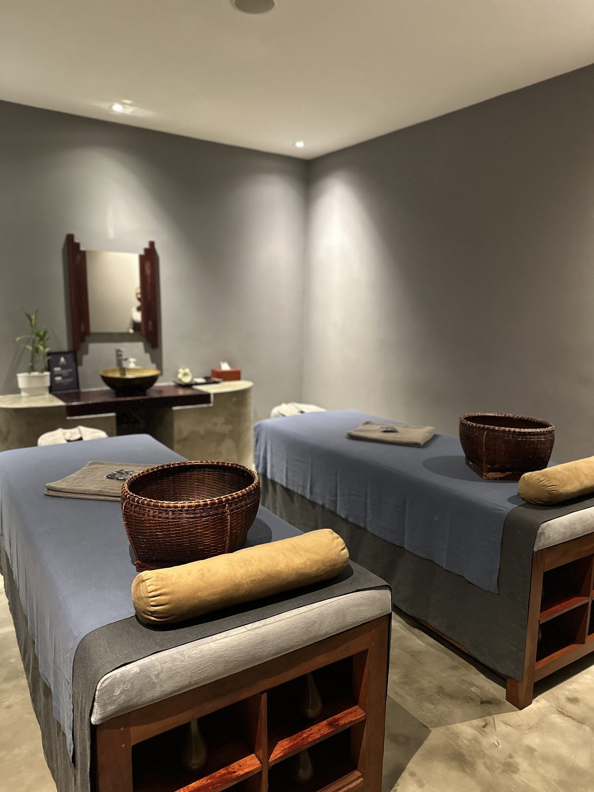 Two massage beds side by side in a treatment room, with bolsters and woven baskets