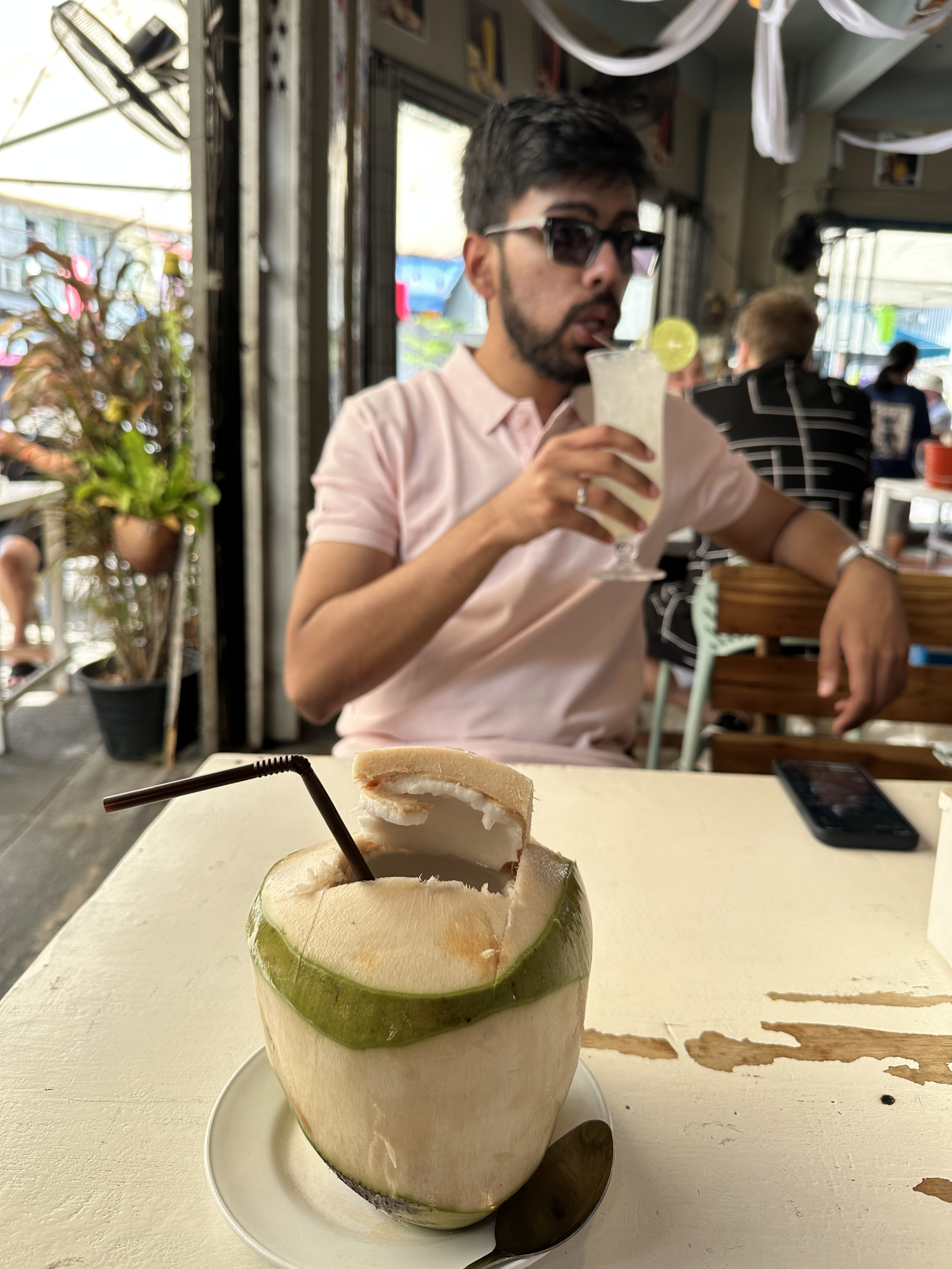 A man drinking from a glass at a Bangkok cafe table with a fresh young coconut in the foreground
