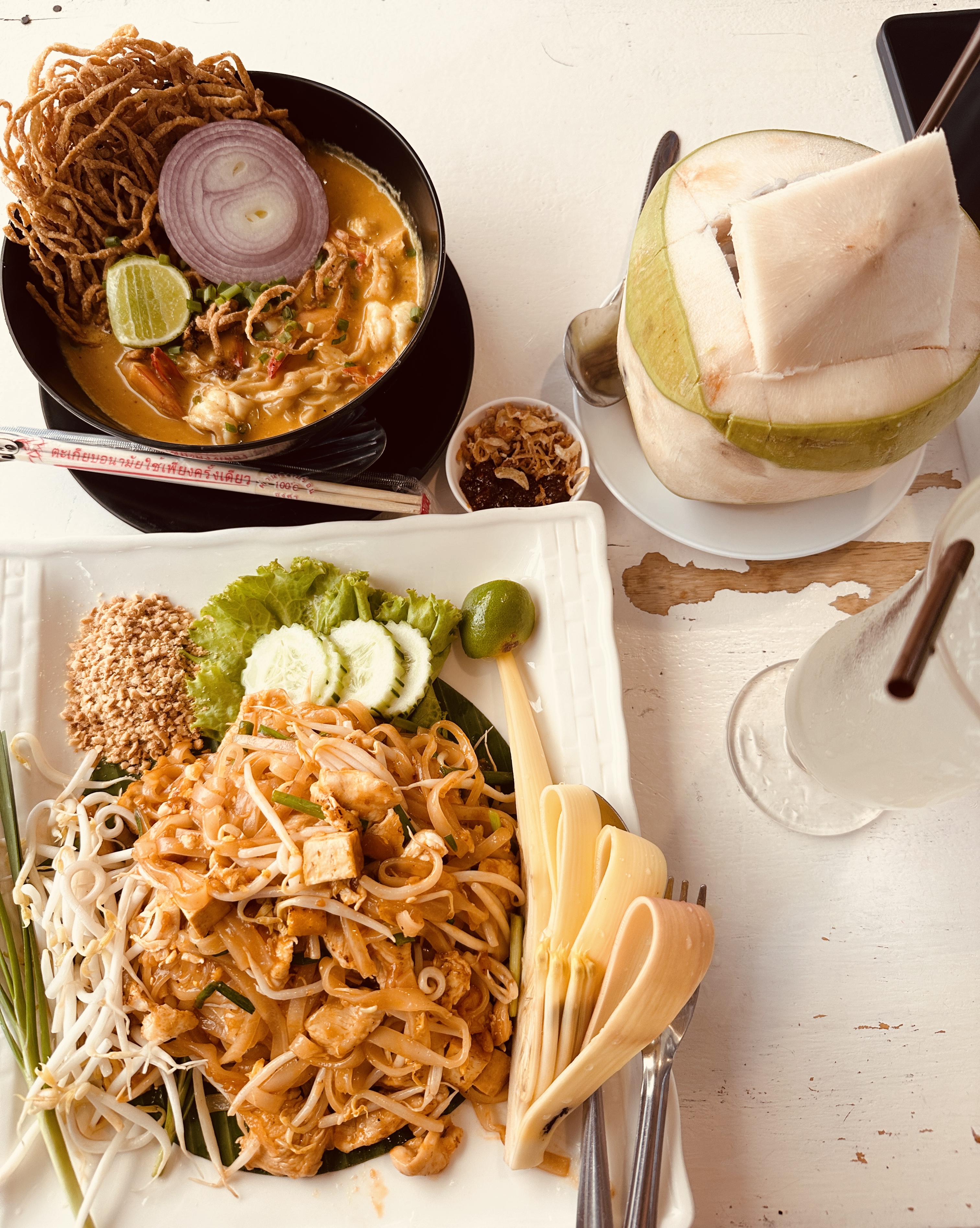 A table of Thai food in Bangkok with pad thai, a bowl of curry noodles and fresh coconuts