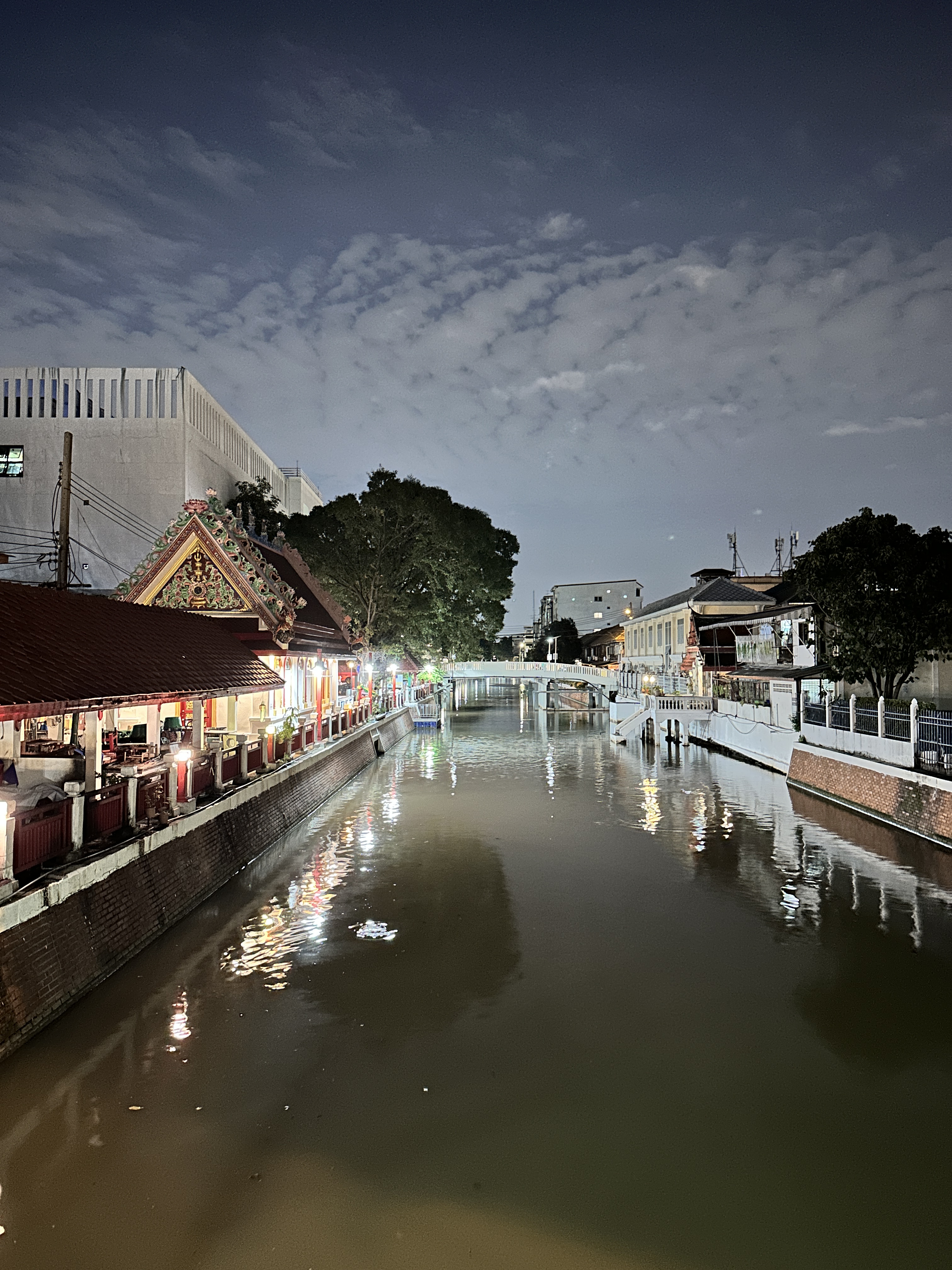 A canal in Bangkok at dusk with a lantern-lit shrine on the left bank and a white footbridge in the distance