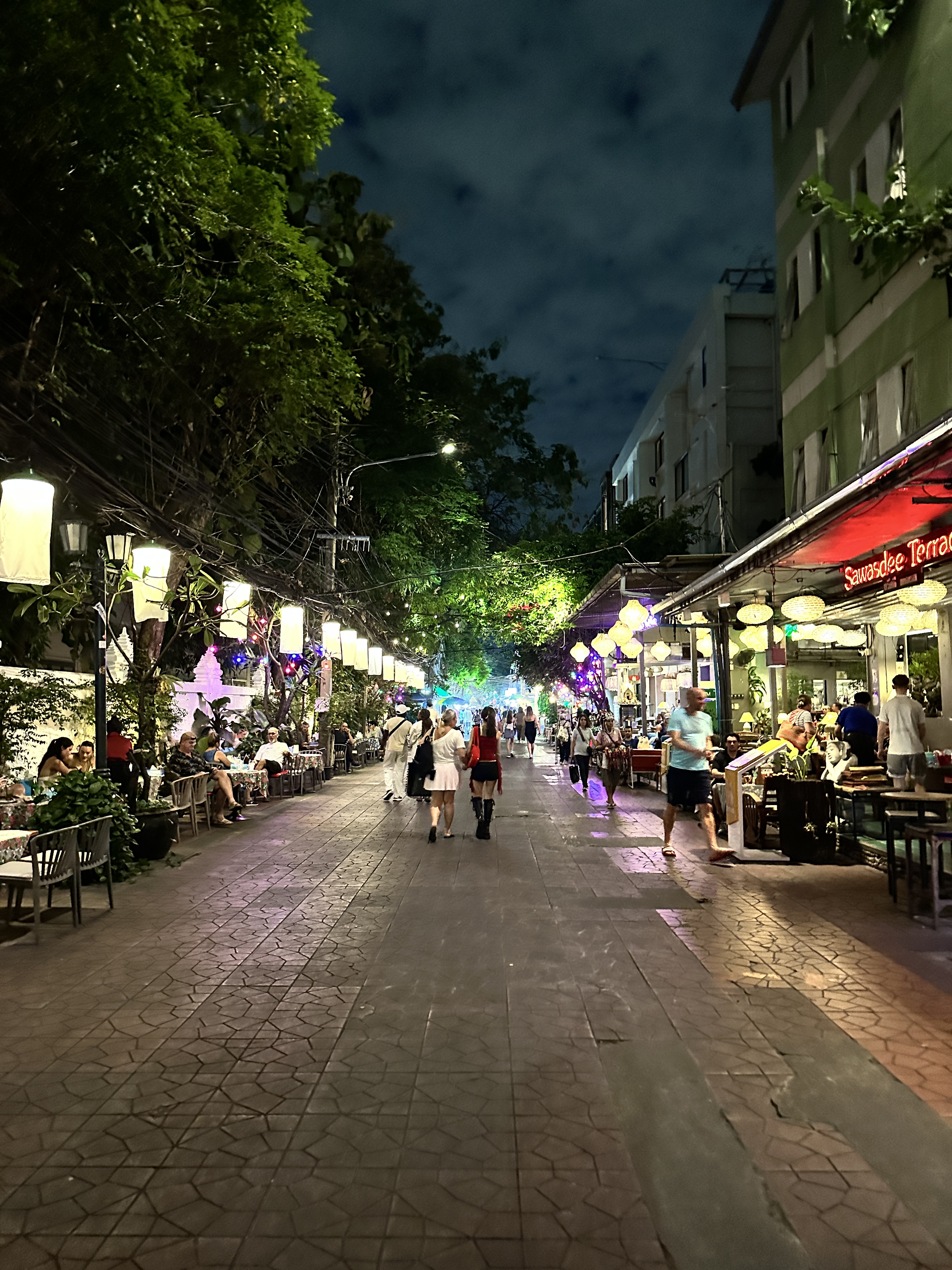 A street in Bangkok at night lined with shops and lights under dark trees