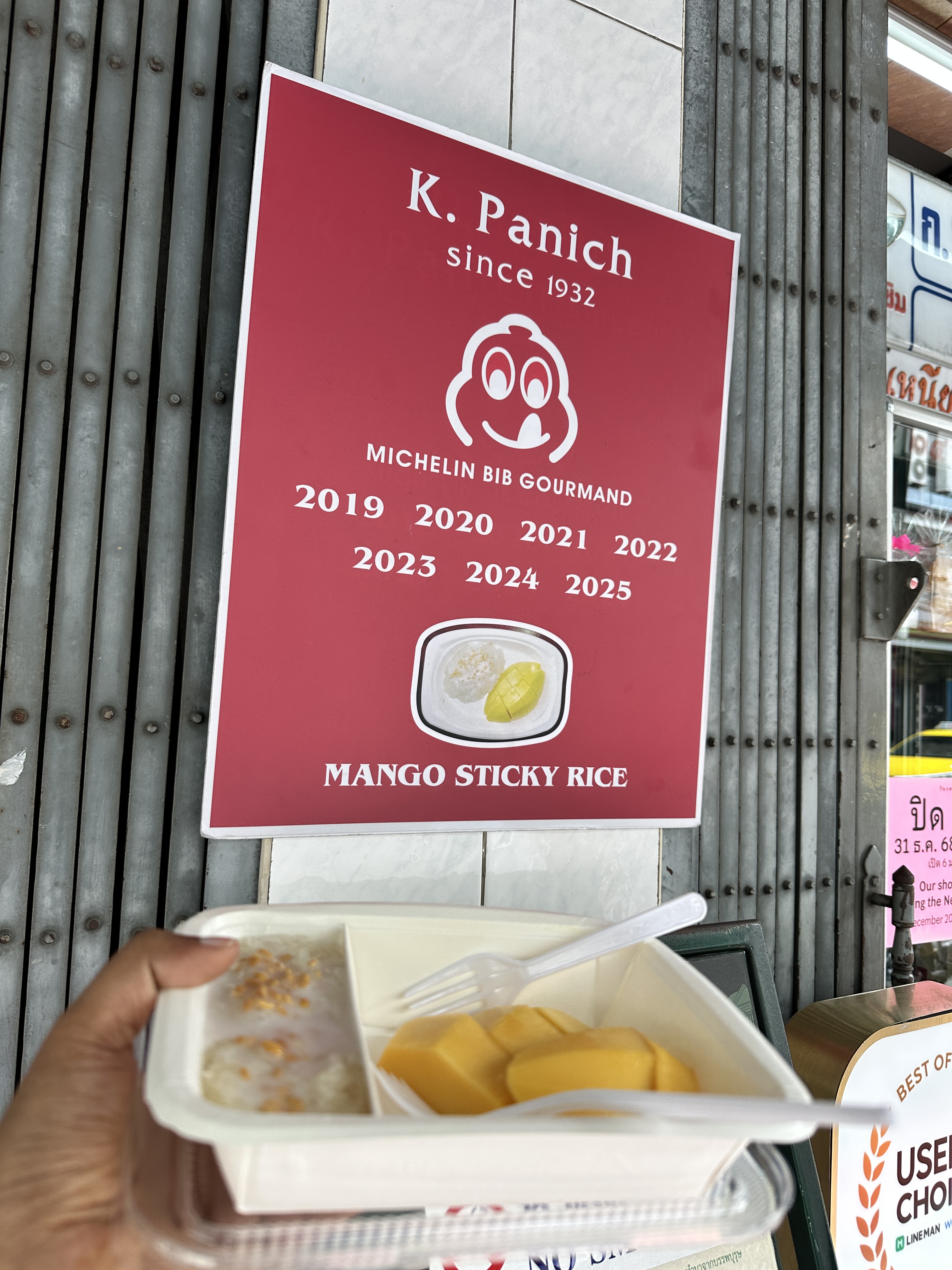 A takeaway box of mango sticky rice held up in front of the K. Panich sign in Bangkok showing Michelin Bib Gourmand awards from 2019 to 2025