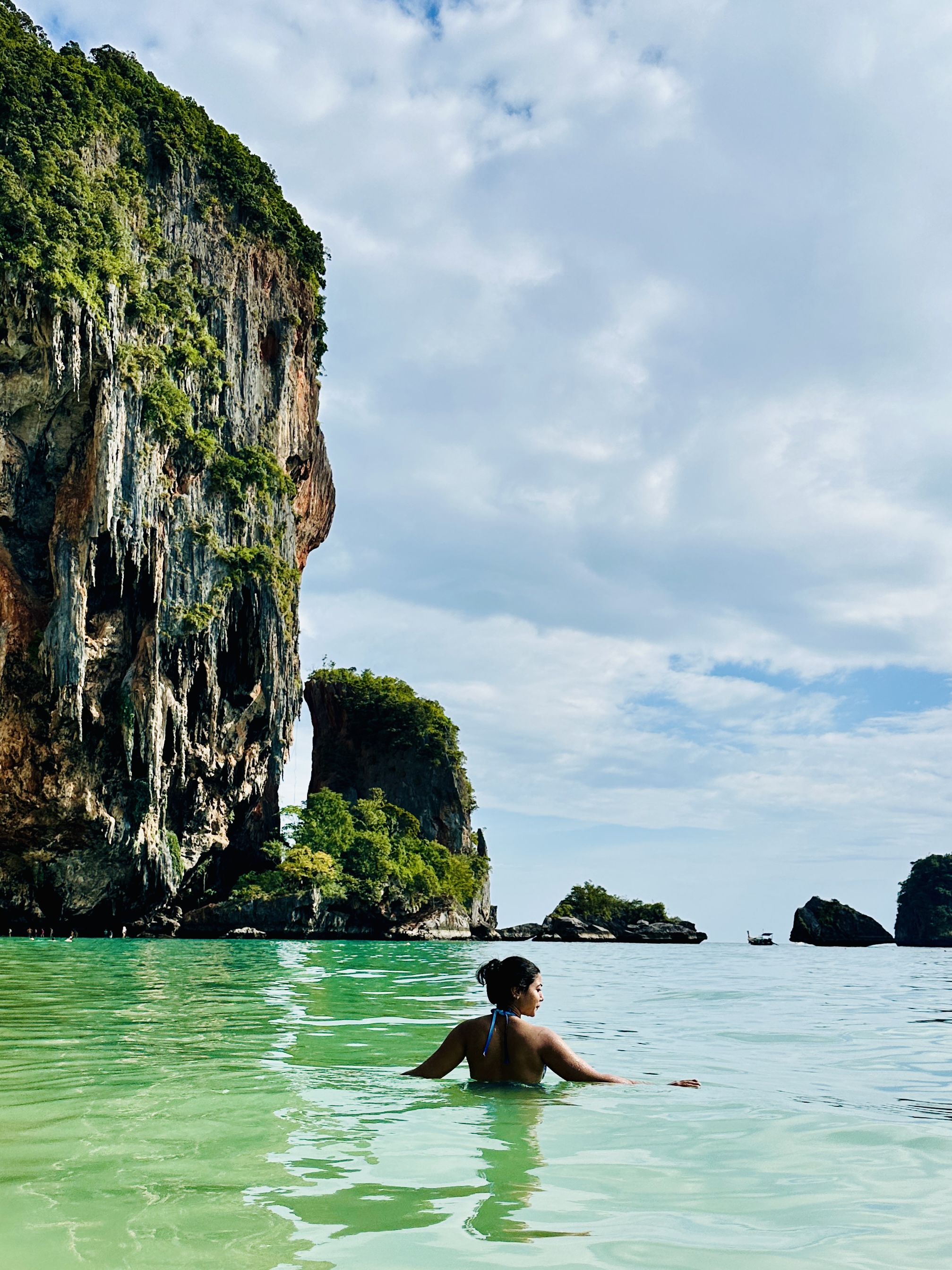 A person swimming in clear green water below a towering limestone cliff at Railay