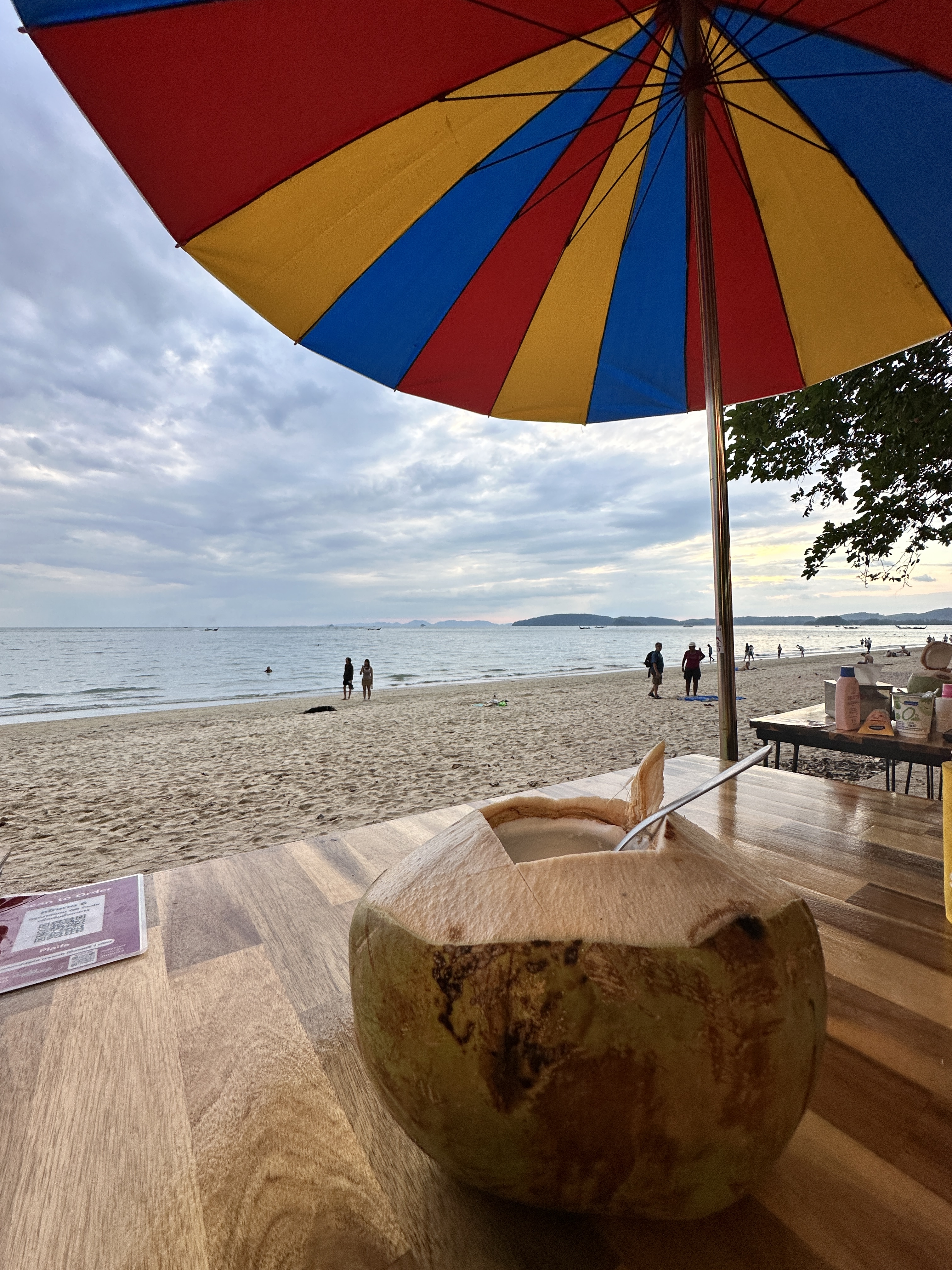 A fresh coconut on a wooden table under a striped beach umbrella looking out to the sea at Railay