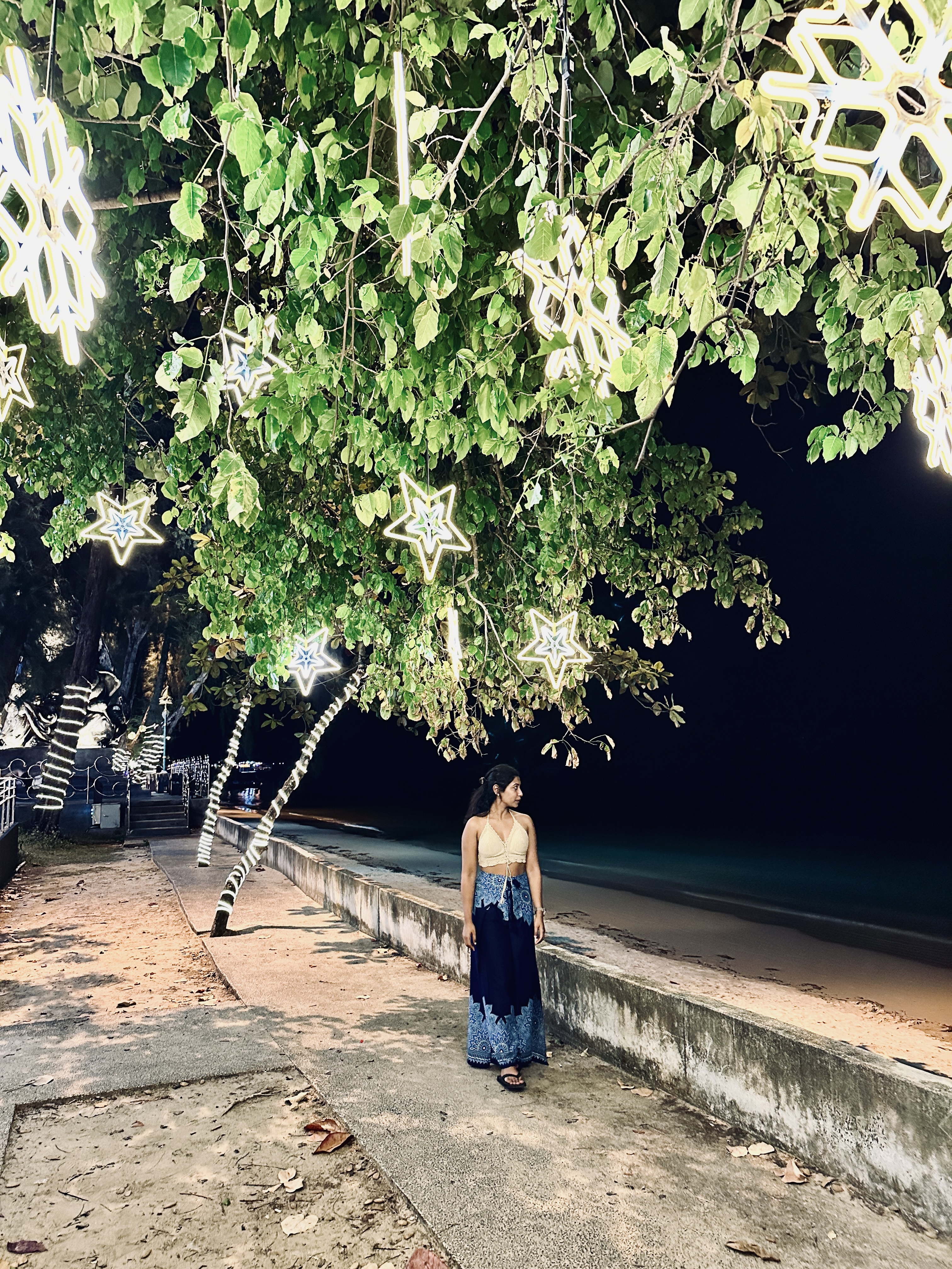 A woman standing on a beachfront path at night beneath trees hung with white paper snowflake decorations