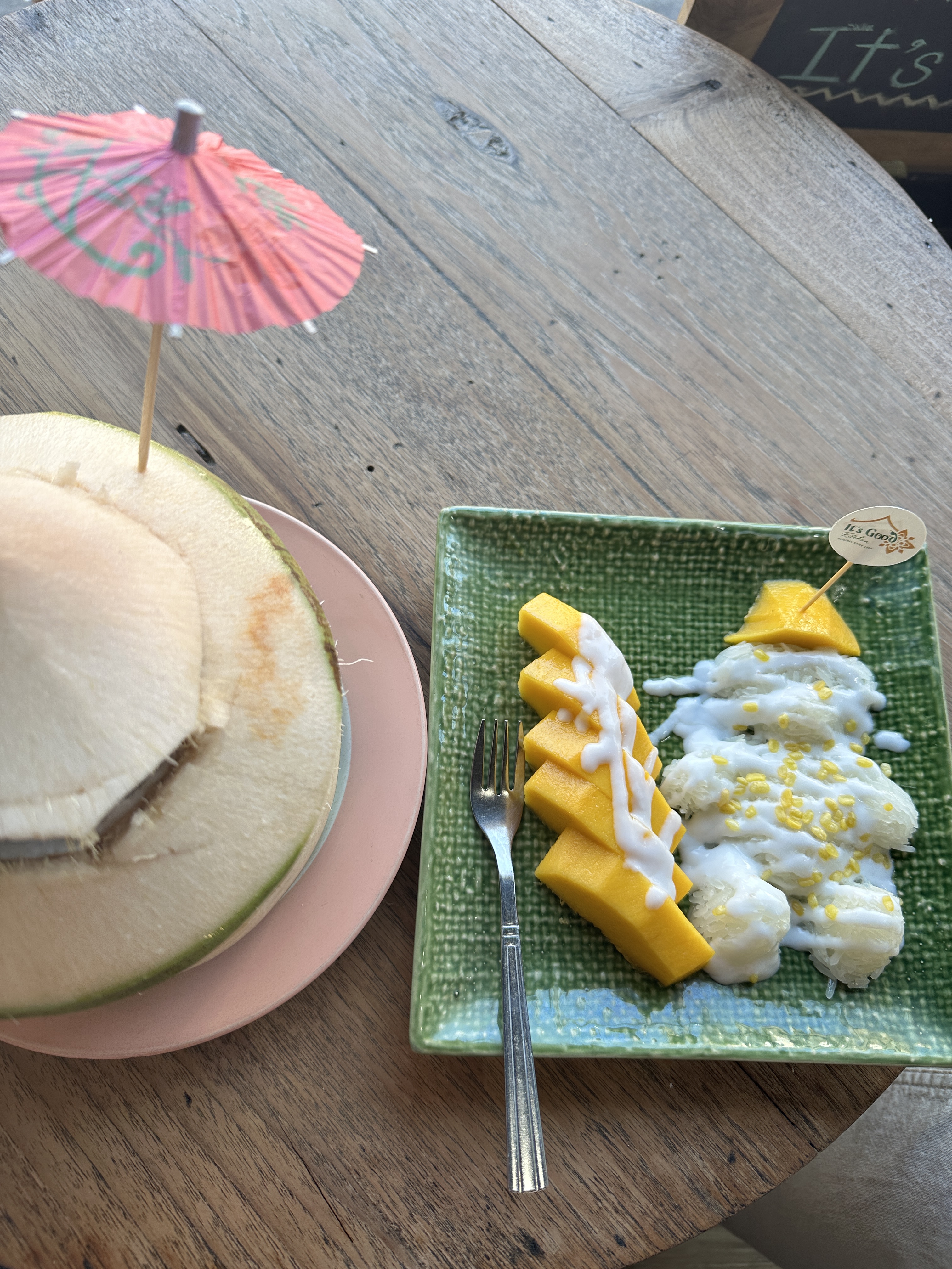 Mango sticky rice on a green plate beside a young coconut with a paper umbrella on a wooden table