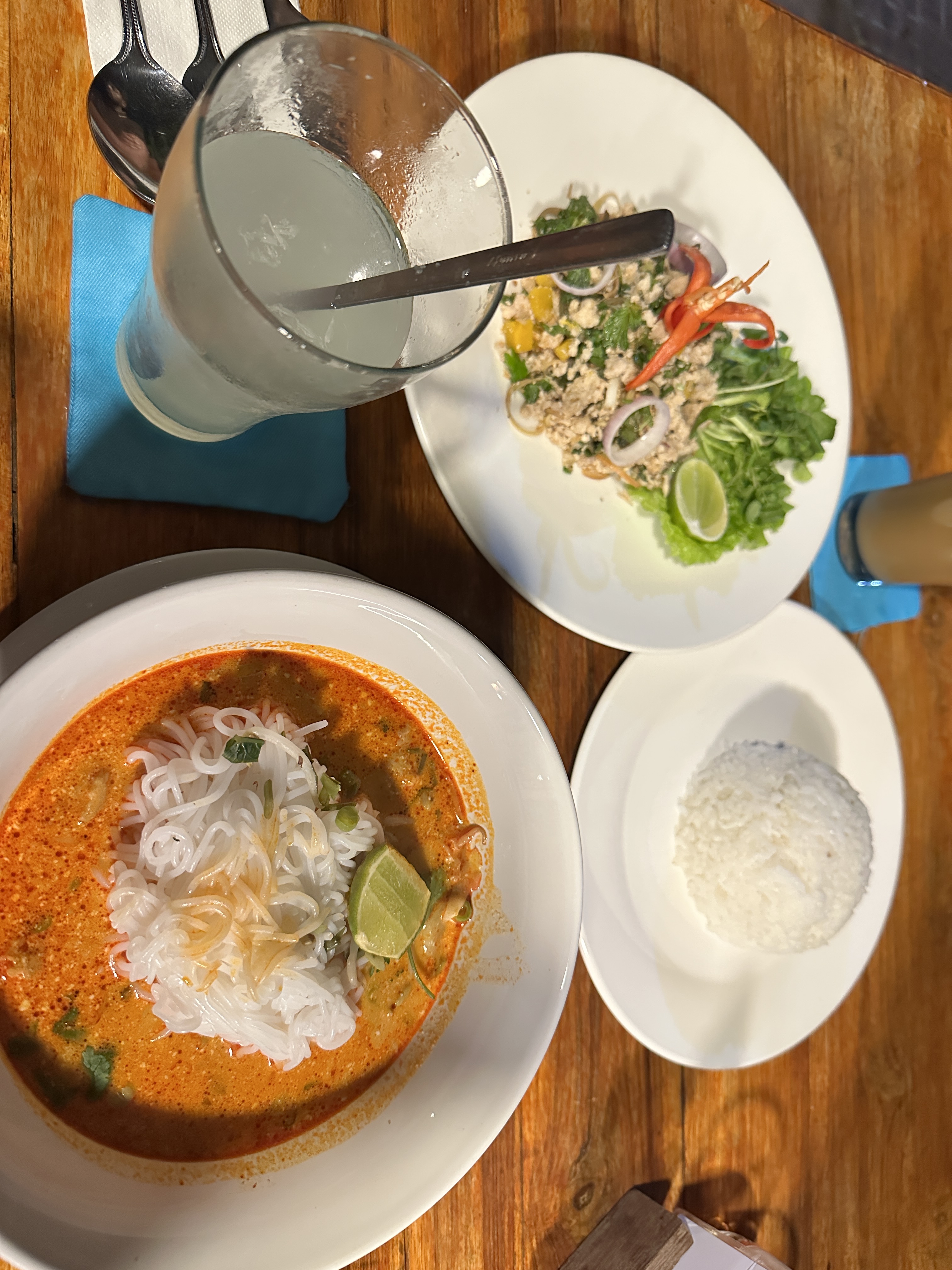 A bowl of khao poon in red coconut broth beside a plate of laap and a bowl of sticky rice