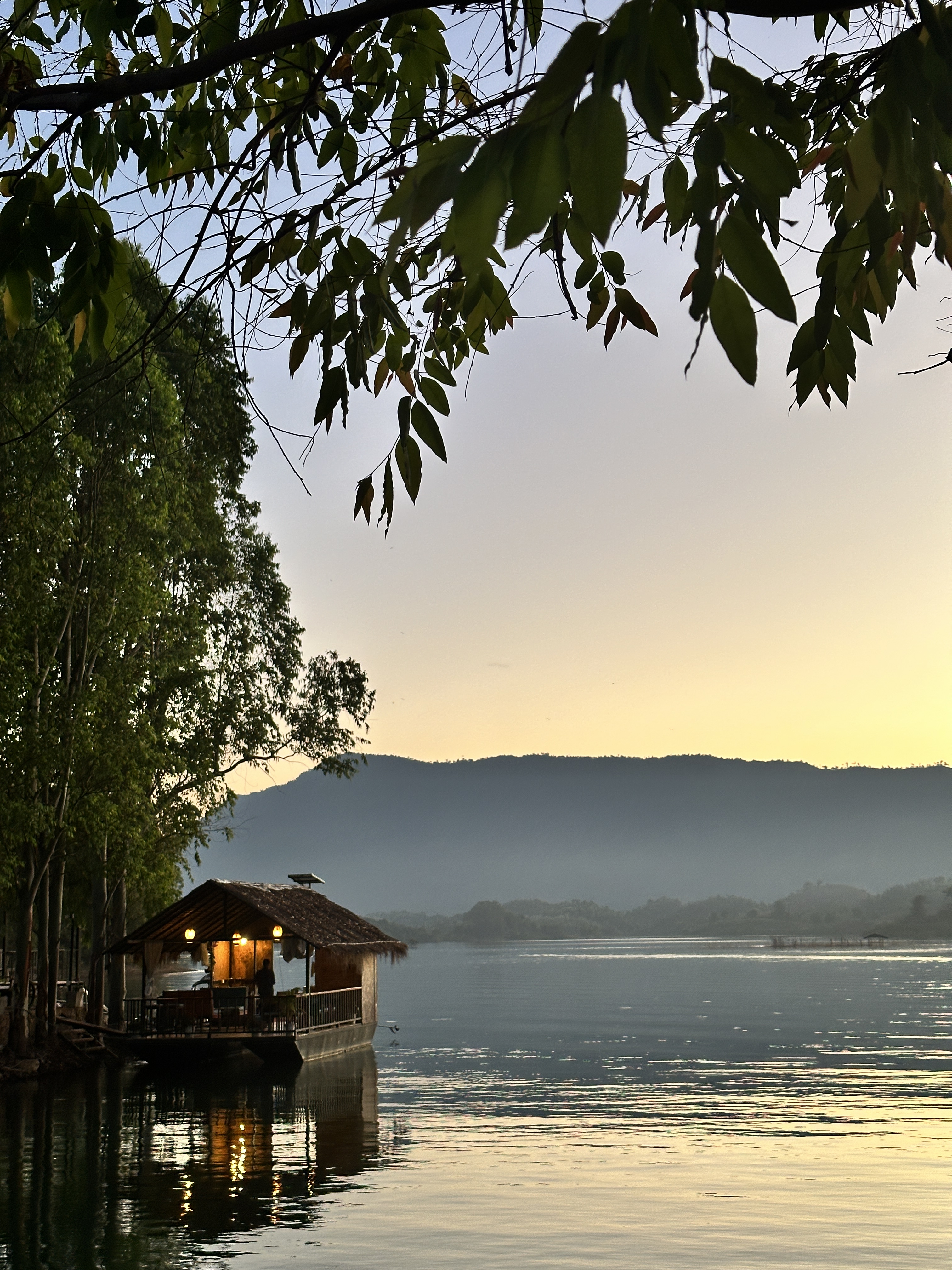 A thatched houseboat on Nam Ngum lake at dusk, lit gold, with hills behind