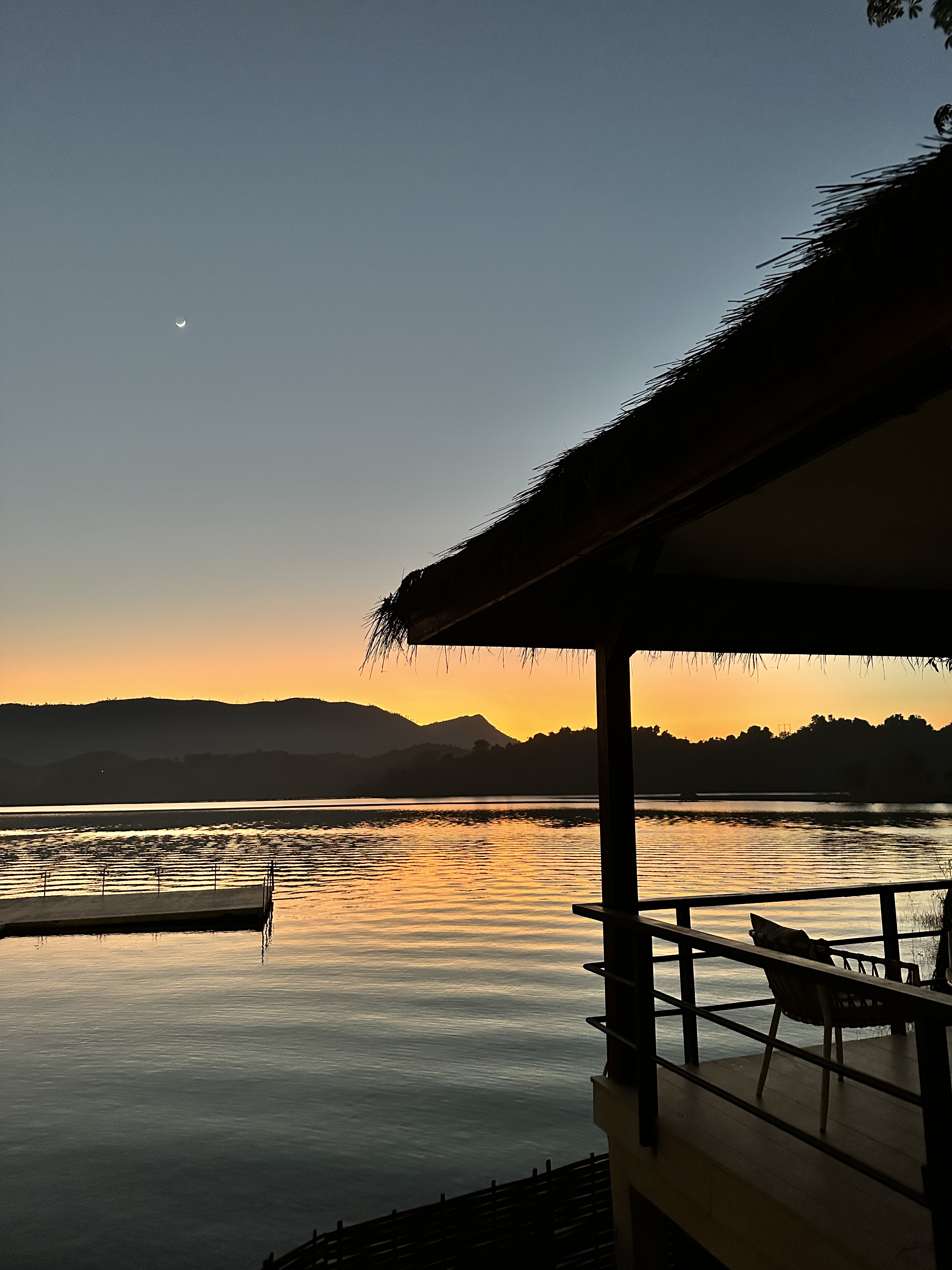 A thin crescent moon over Nam Ngum lake at sunset, beside a thatched pavilion