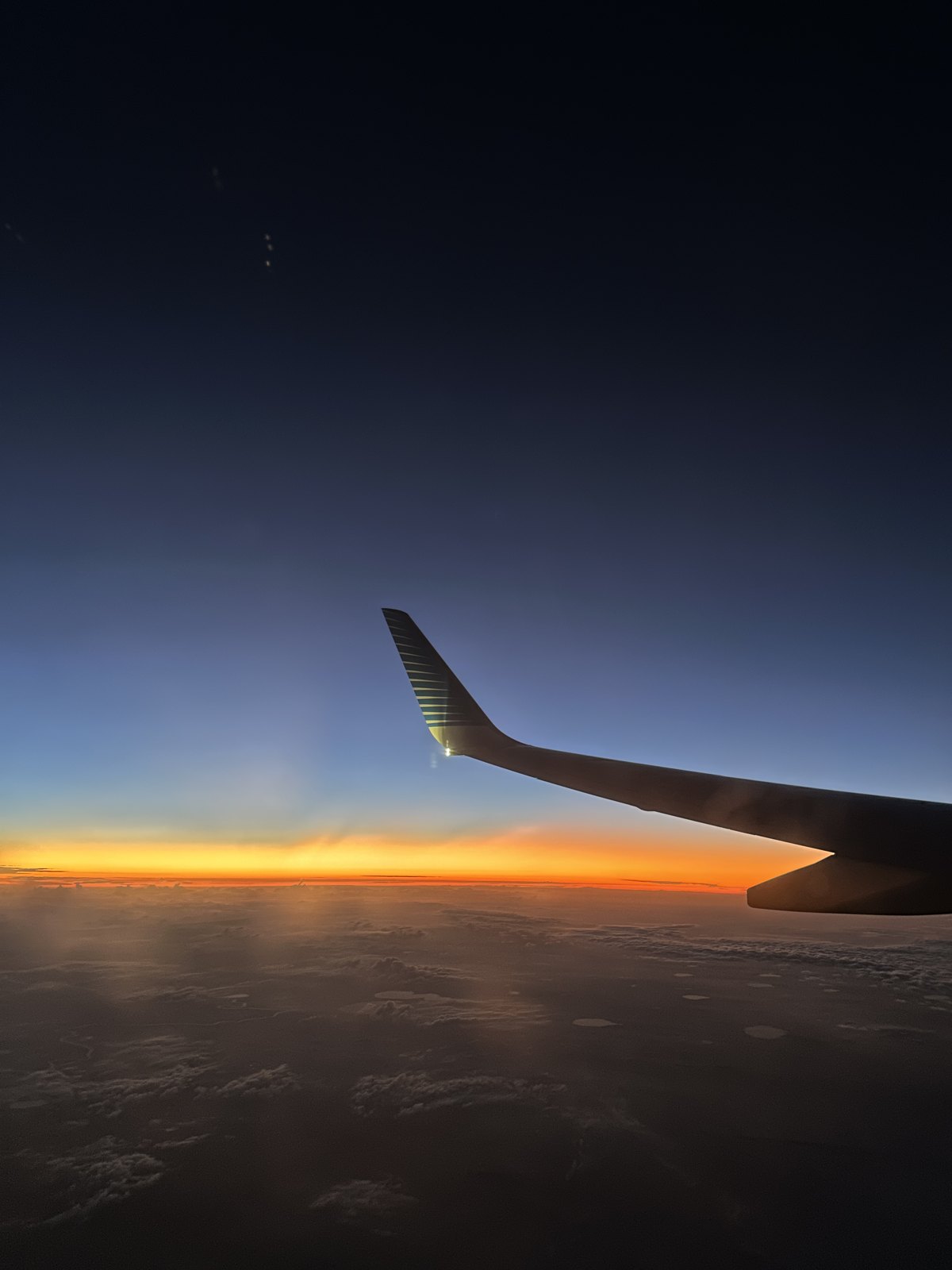 Sunrise seen from an aircraft window, the wing silhouetted against an orange band of light above cloud