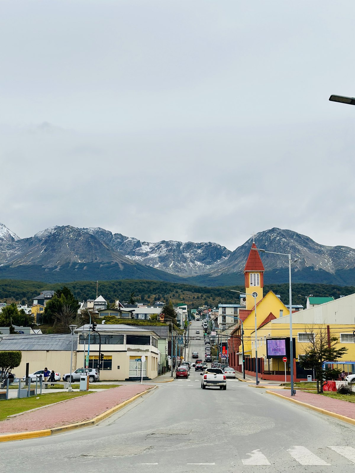 A street in Ushuaia running downhill towards the water, with colourful low buildings and snow-streaked mountains rising behind the town