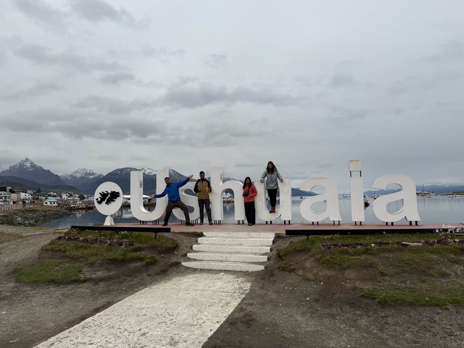 A couple standing among the large white letters spelling USHUAIA on the waterfront, with the harbour and mountains behind