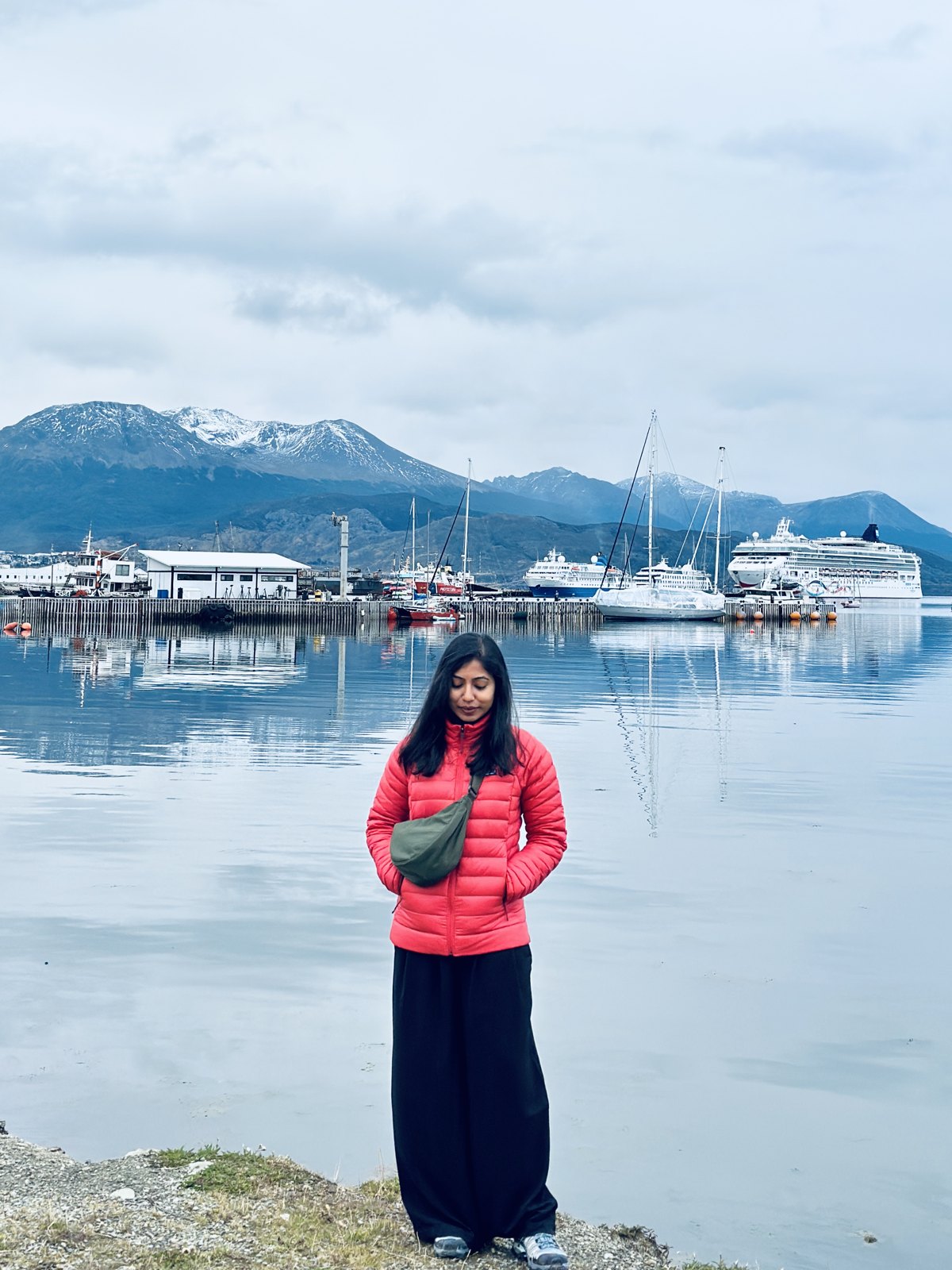 A woman in a red down jacket standing by the still water of Ushuaia harbour, with yachts, a cruise ship and snow-capped mountains reflected behind her