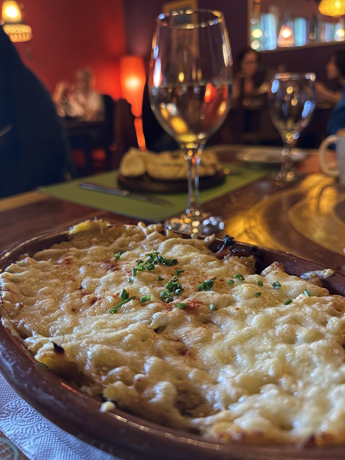 A bubbling baked dish covered in melted cheese on a dark restaurant table, with a glass of white wine behind it