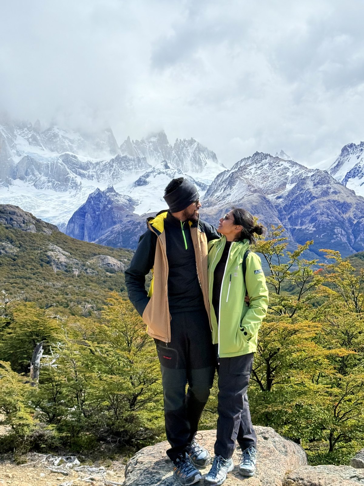 A couple standing on a rock in bright jackets, with the snow-covered granite spires of the Fitz Roy massif rising into cloud behind them