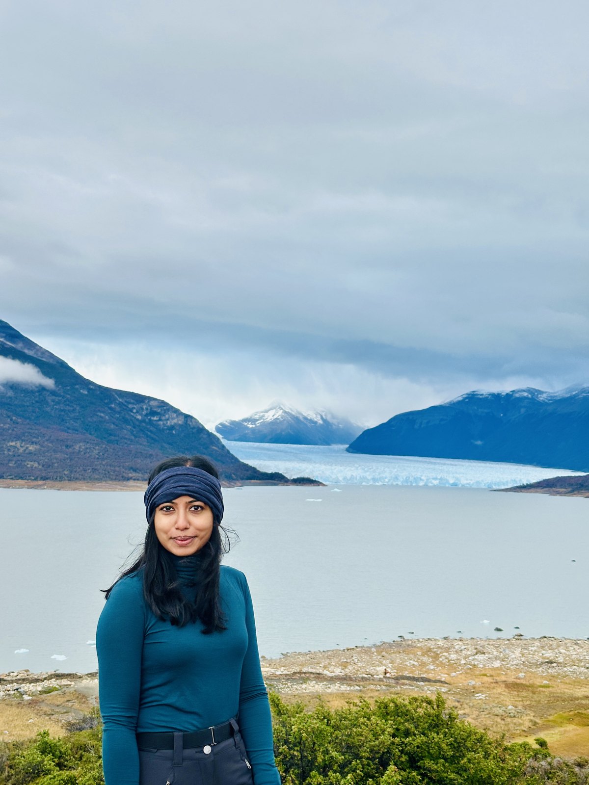 A woman in a teal top and headband standing on a hillside with the pale blue face of the Perito Moreno glacier stretching across the water behind her