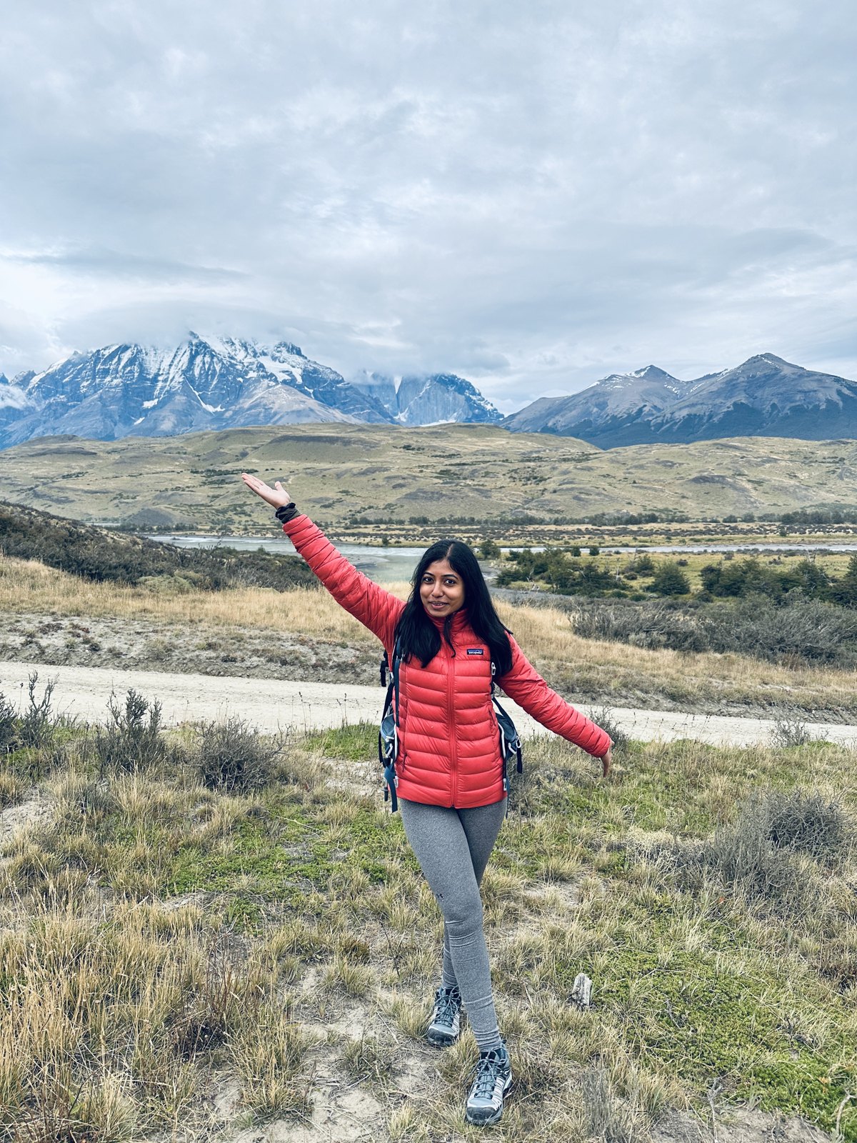 A woman in a red jacket with a pack standing on the steppe with one arm raised, the jagged peaks of the Paine massif behind her