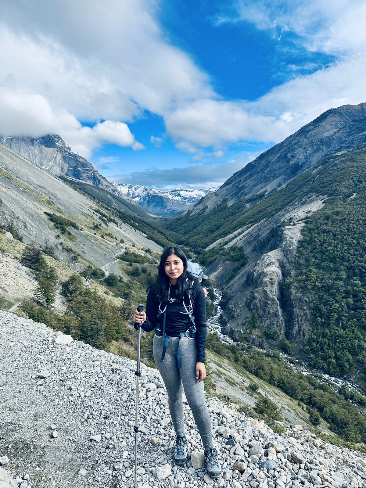 A woman with a trekking pole and a large pack standing on a stony path high above a green glacial valley with snow peaks at its head